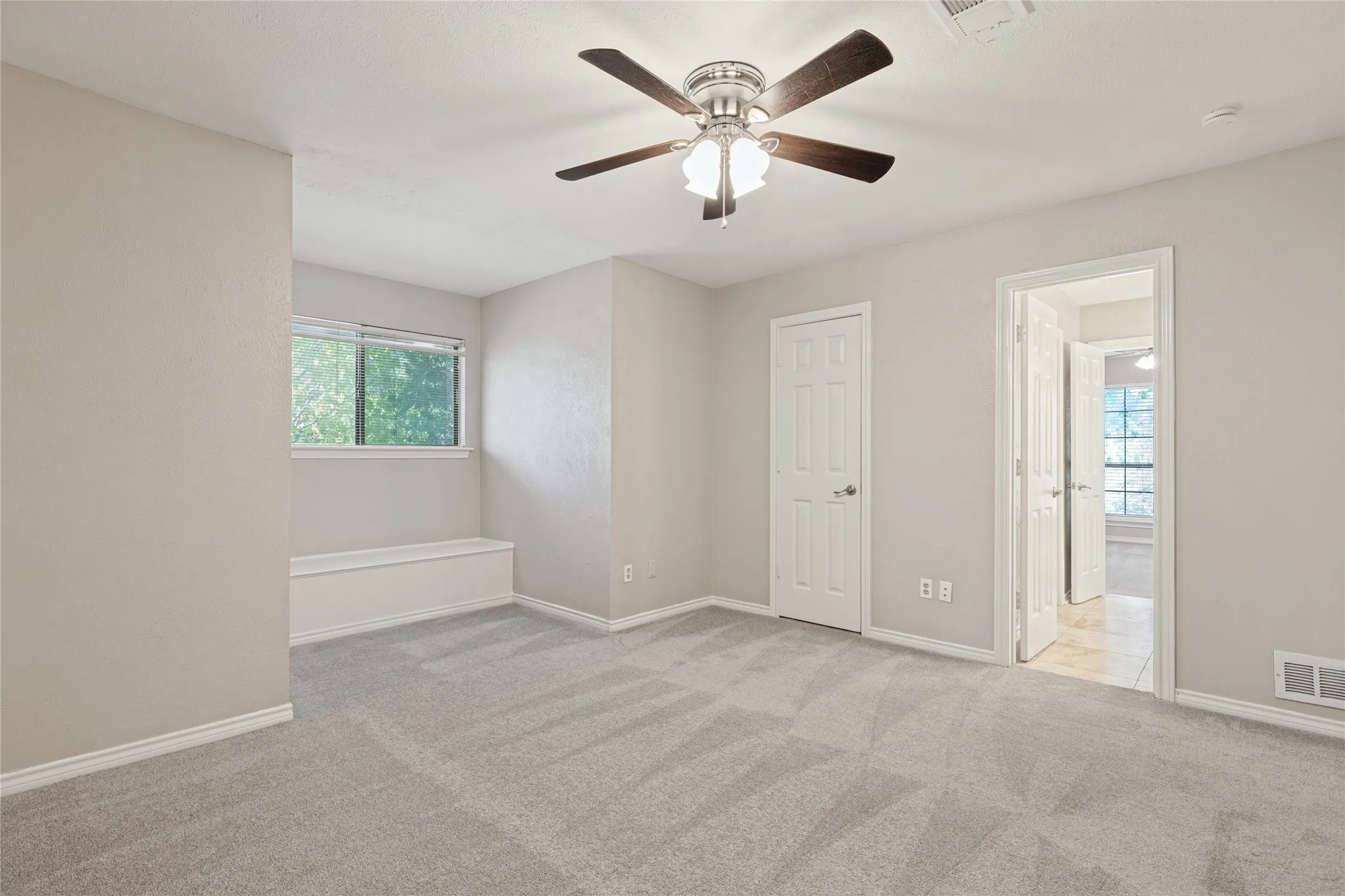 Spacious upstairs bedroom with light neutral wall tones, gray carpeting, a built-in window bench, and a ceiling fan with integrated lighting, attached Jack & Jill Bath