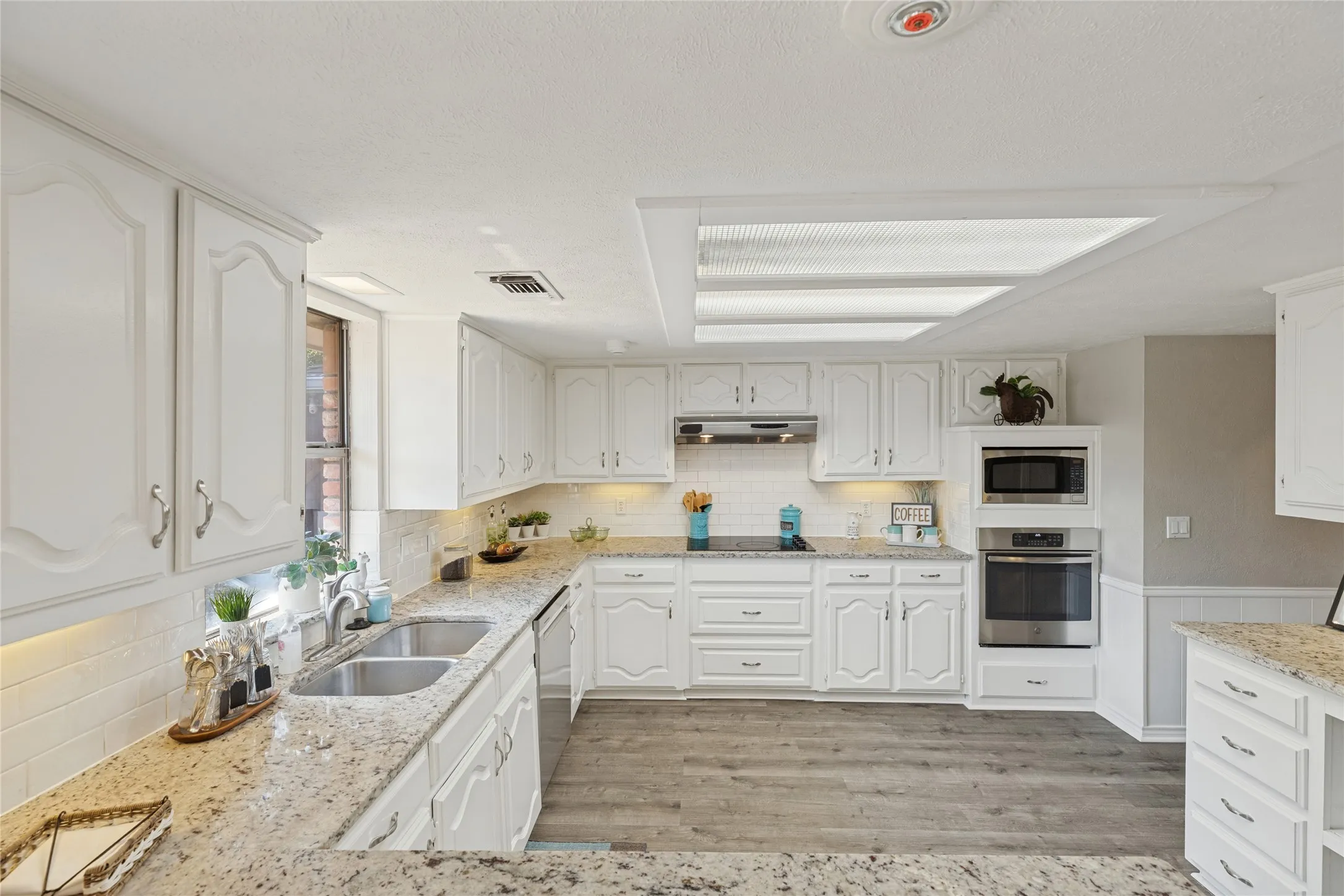 Kitchen featuring white cabinetry, light-toned countertops, a double basin sink, stainless steel built-in appliances, and wood-finish flooring, granite countertops
