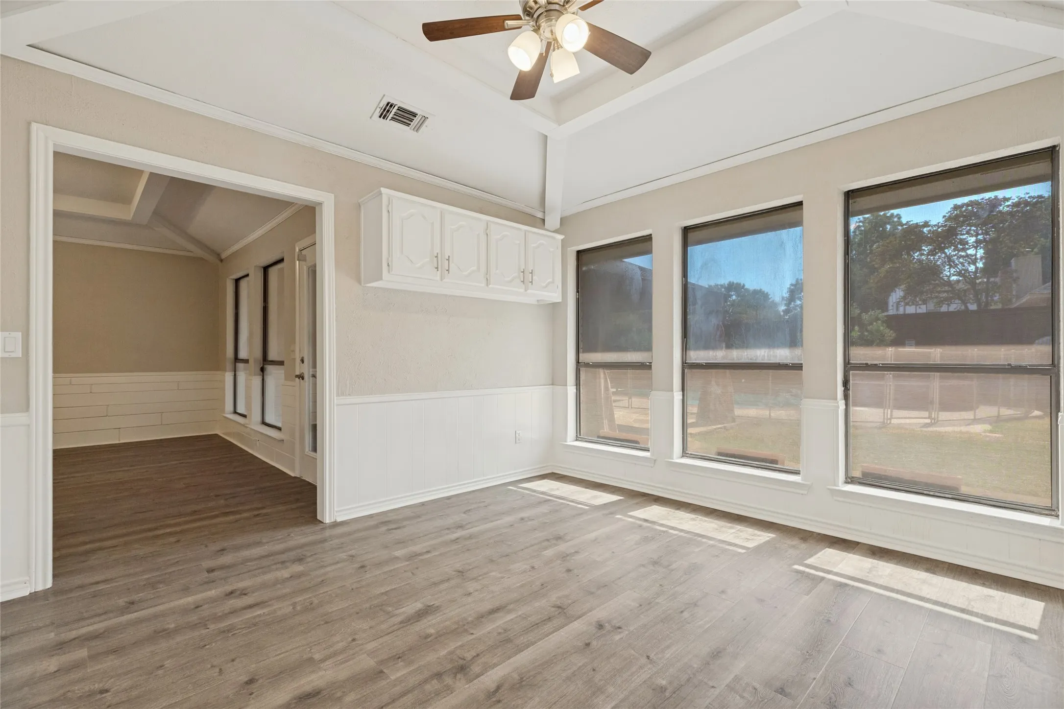 Vaulted ceiling with a ceiling fan, wood-finish flooring, three large windows, white cabinetry, and wainscoting