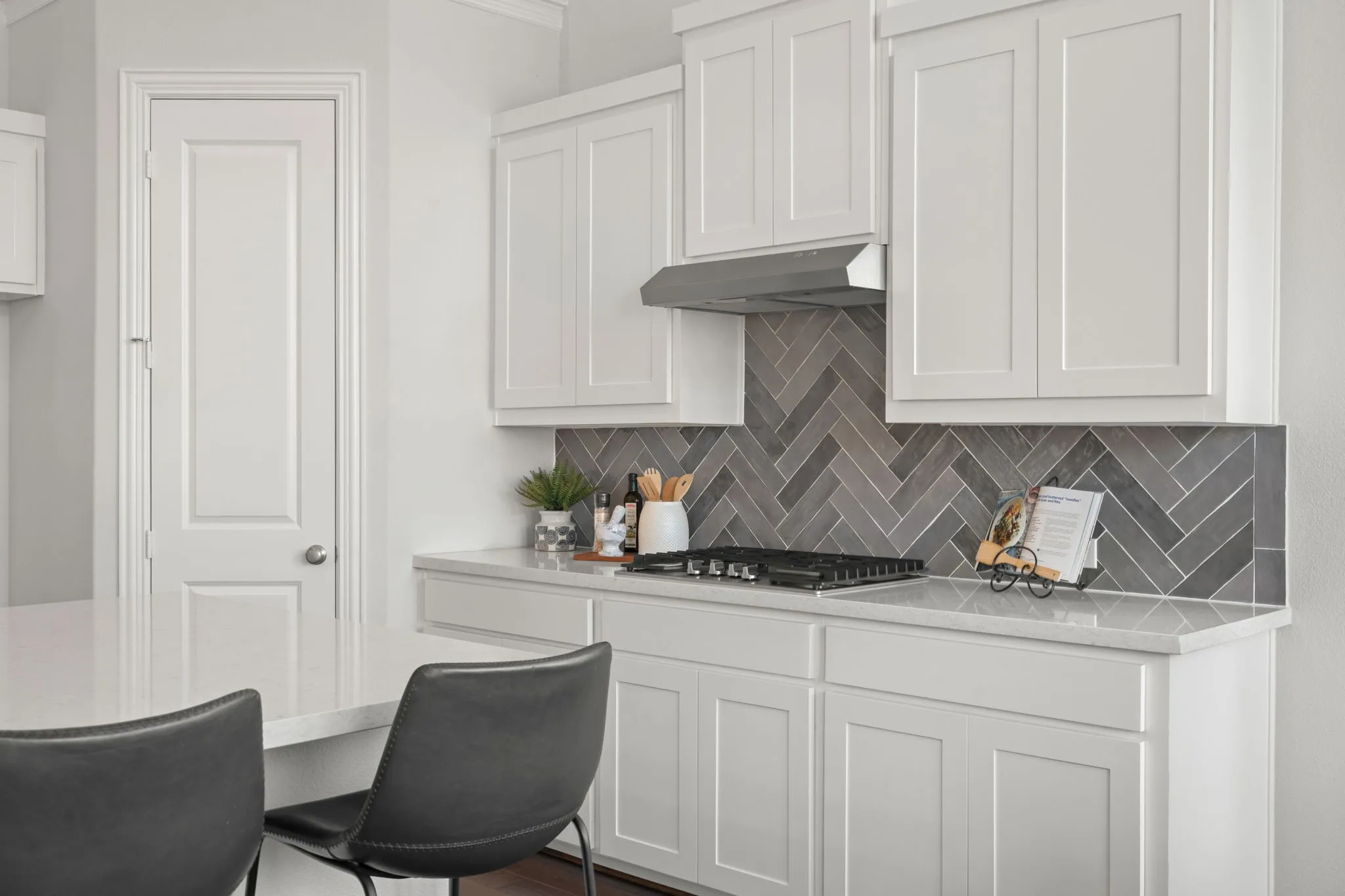 White shaker cabinetry, light-toned countertops, herringbone tile backsplash, stainless steel range hood, and a built-in gas cooktop