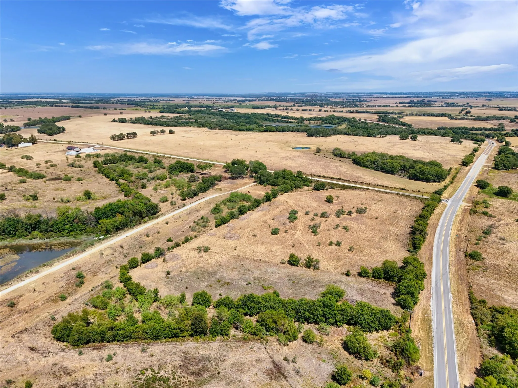 Expansive open land featuring a paved road, scattered mature trees, and a visible pond