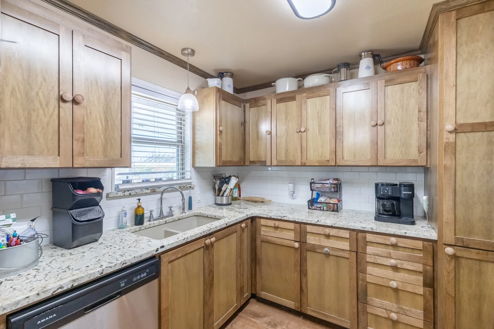 Kitchen featuring wood cabinetry, light-toned countertops, subway tile backsplash, stainless steel dishwasher, and a double basin sink