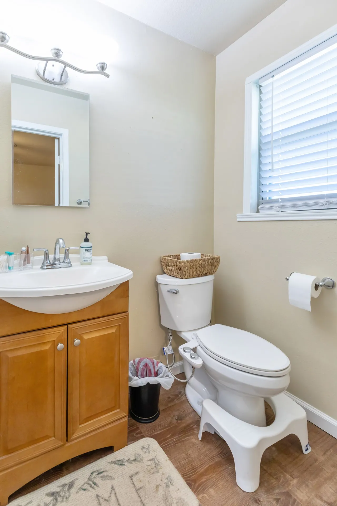 Wood-finish flooring, single vanity with wood cabinetry, window with blinds, wall-mounted mirror, and a modern toilet
