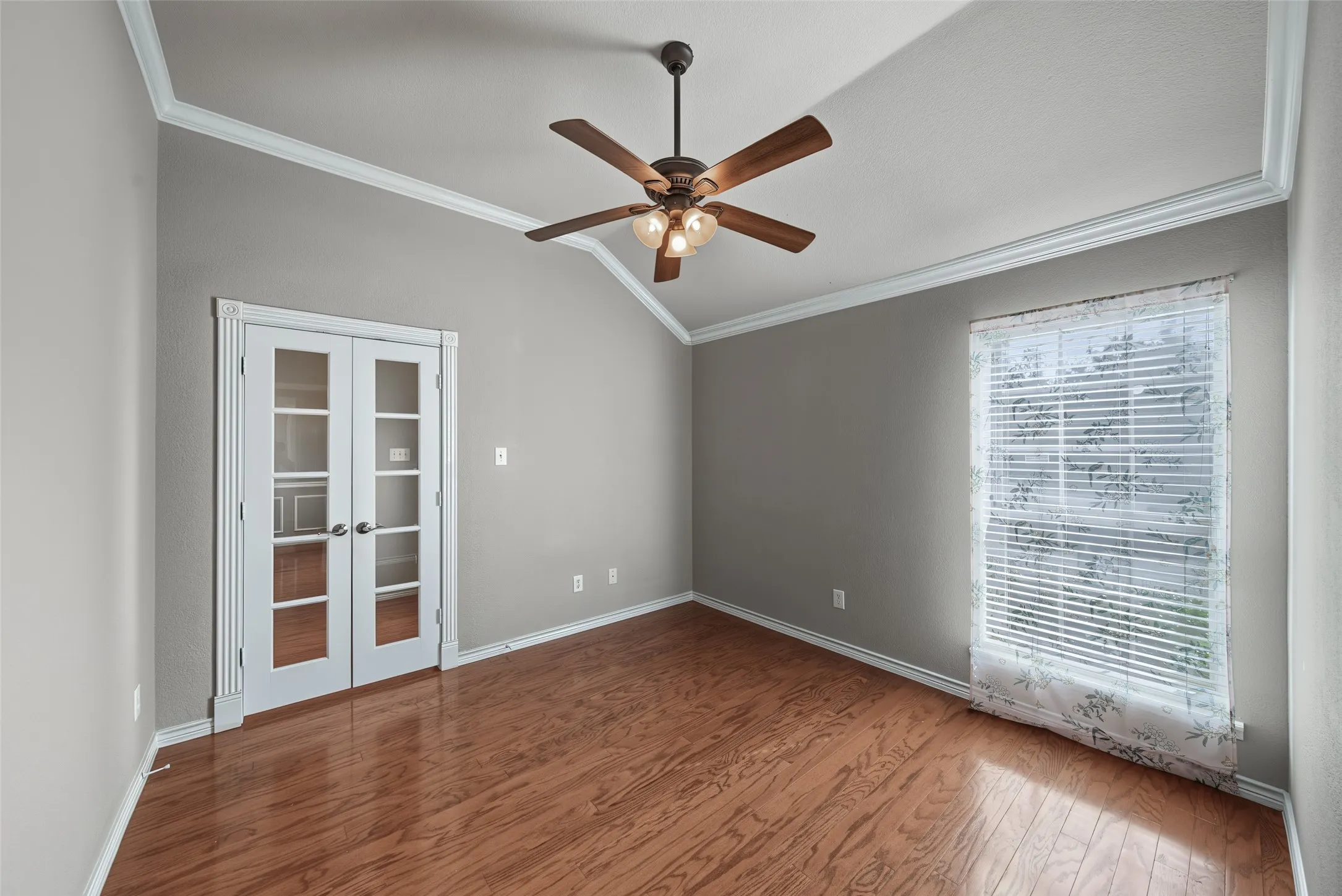 Vaulted ceiling with crown molding, wood-finish flooring, French doors with glass panels, ceiling fan with integrated lighting, and a large window with blinds