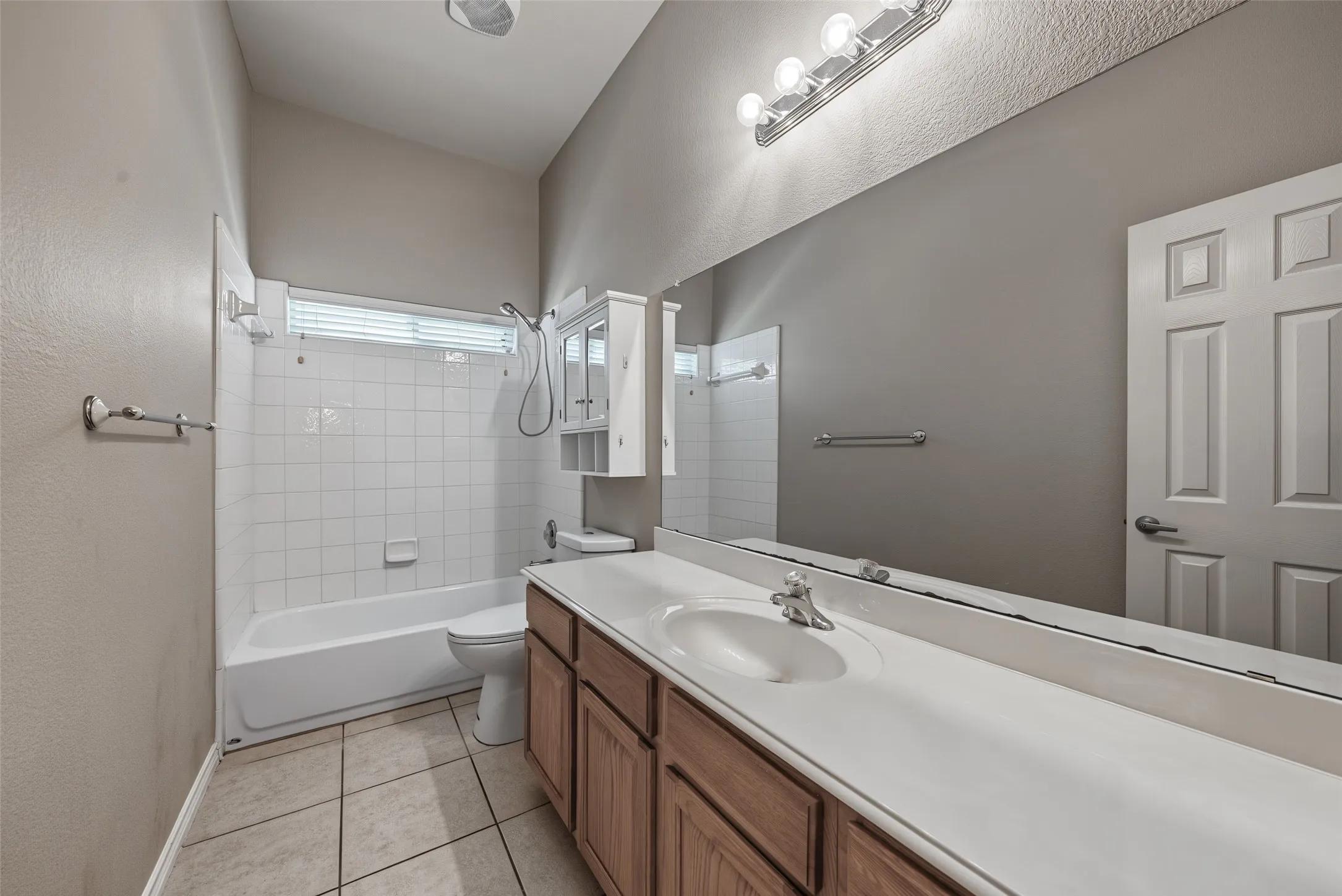 Bathroom featuring a single vanity with wood-finish cabinetry, a large integrated countertop and sink, a full-width mirror, and a white tile shower-tub combination with an overhead window