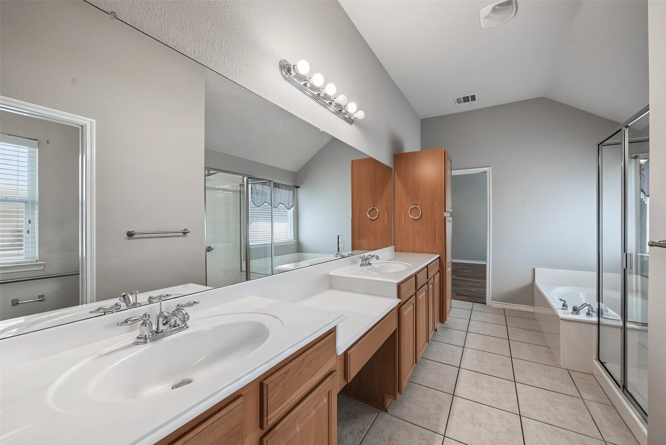 Dual vanity with integrated sinks, wood-finish cabinetry, tile flooring, separate shower enclosure, and soaking tub