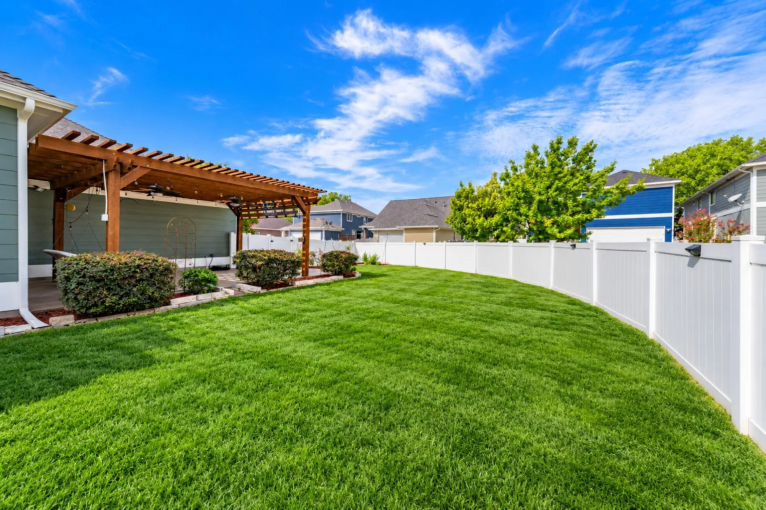 Spacious backyard featuring a wooden pergola, concrete patio, privacy fencing, manicured lawn, and planted shrubs