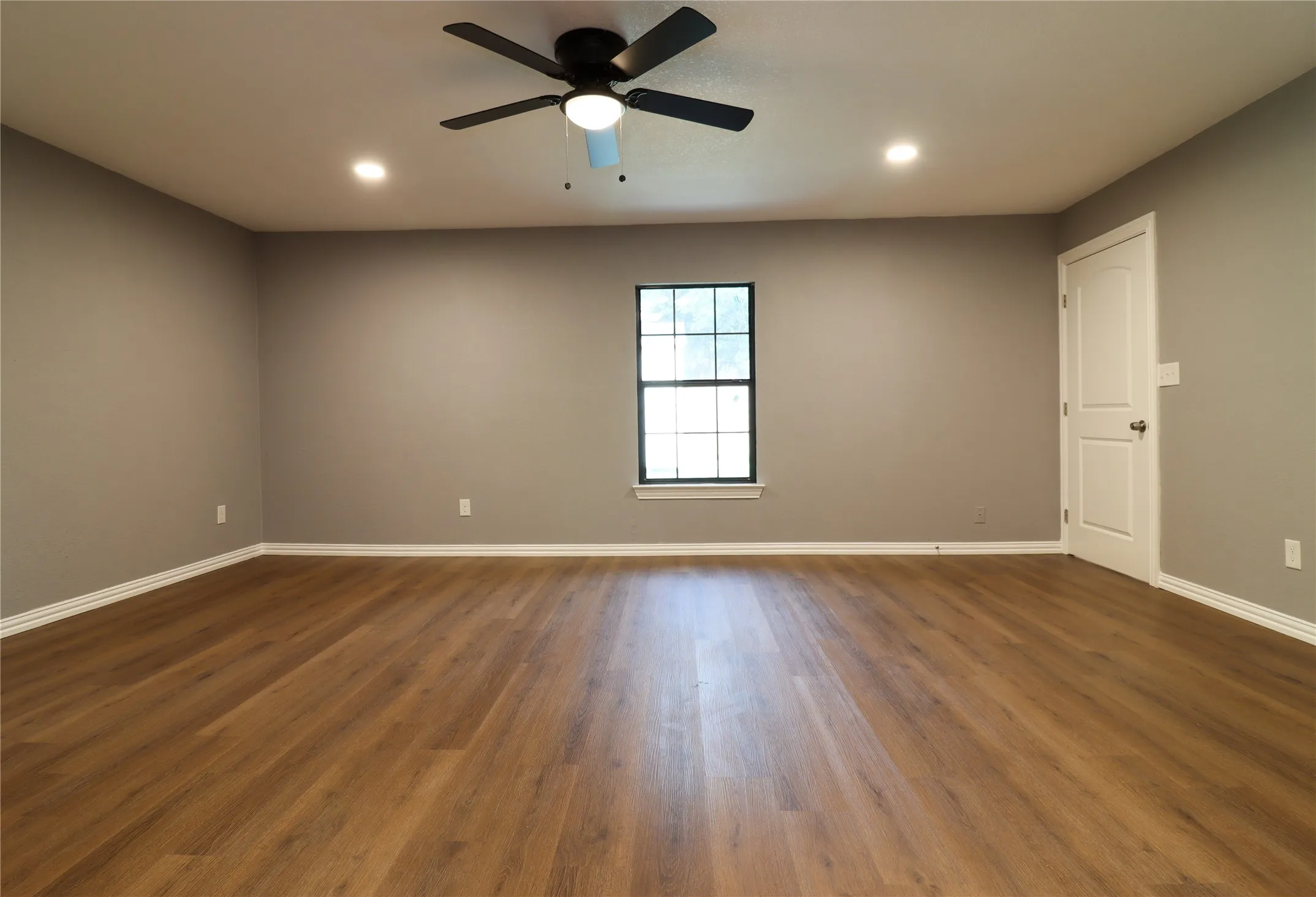 Spacious room featuring gleaming wood-finish flooring, neutral wall finishes, and abundant natural light from a large fixed window