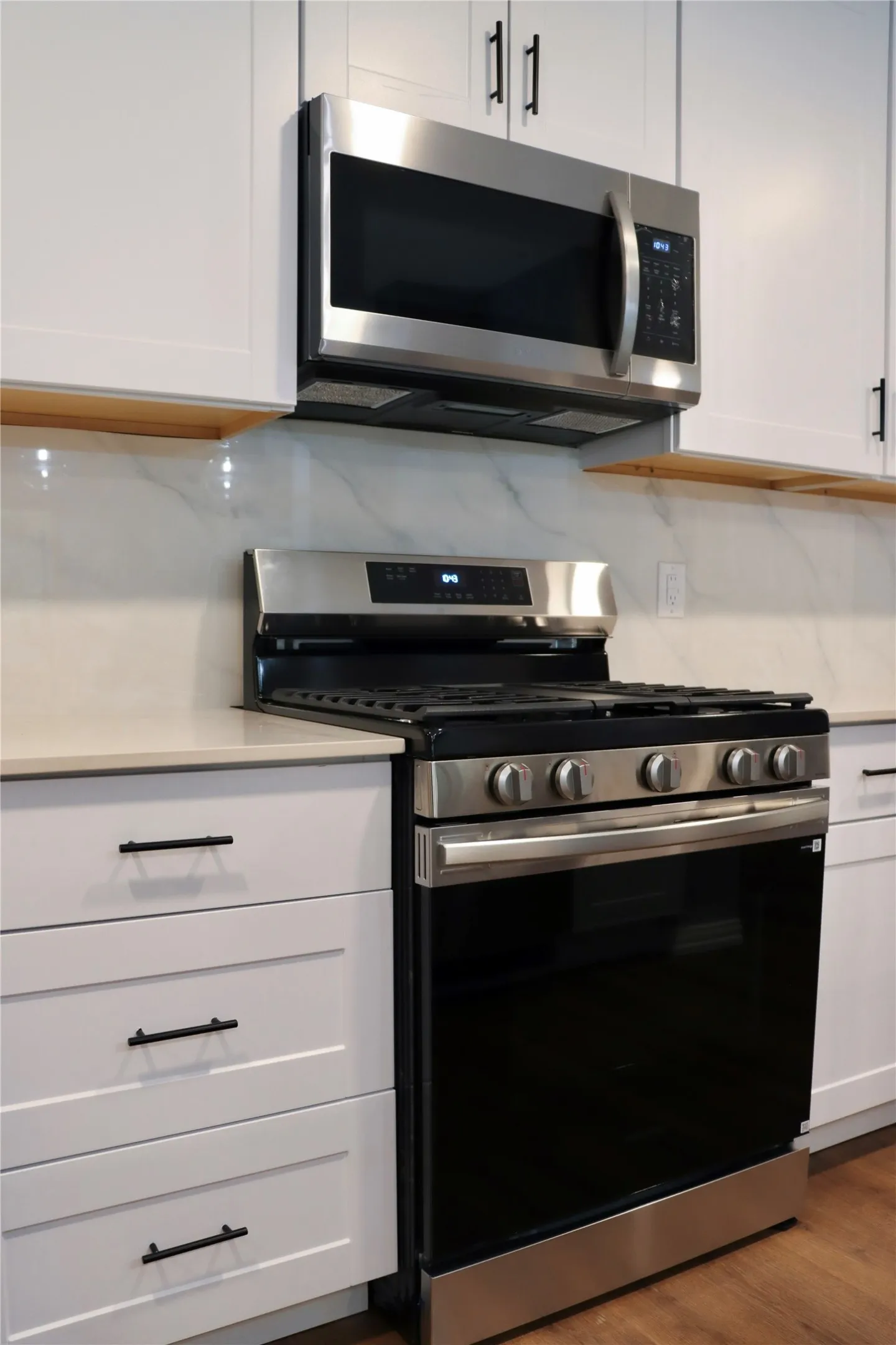 Stainless steel microwave and range, white shaker-style cabinetry, light-toned countertop, and wood-finish flooring