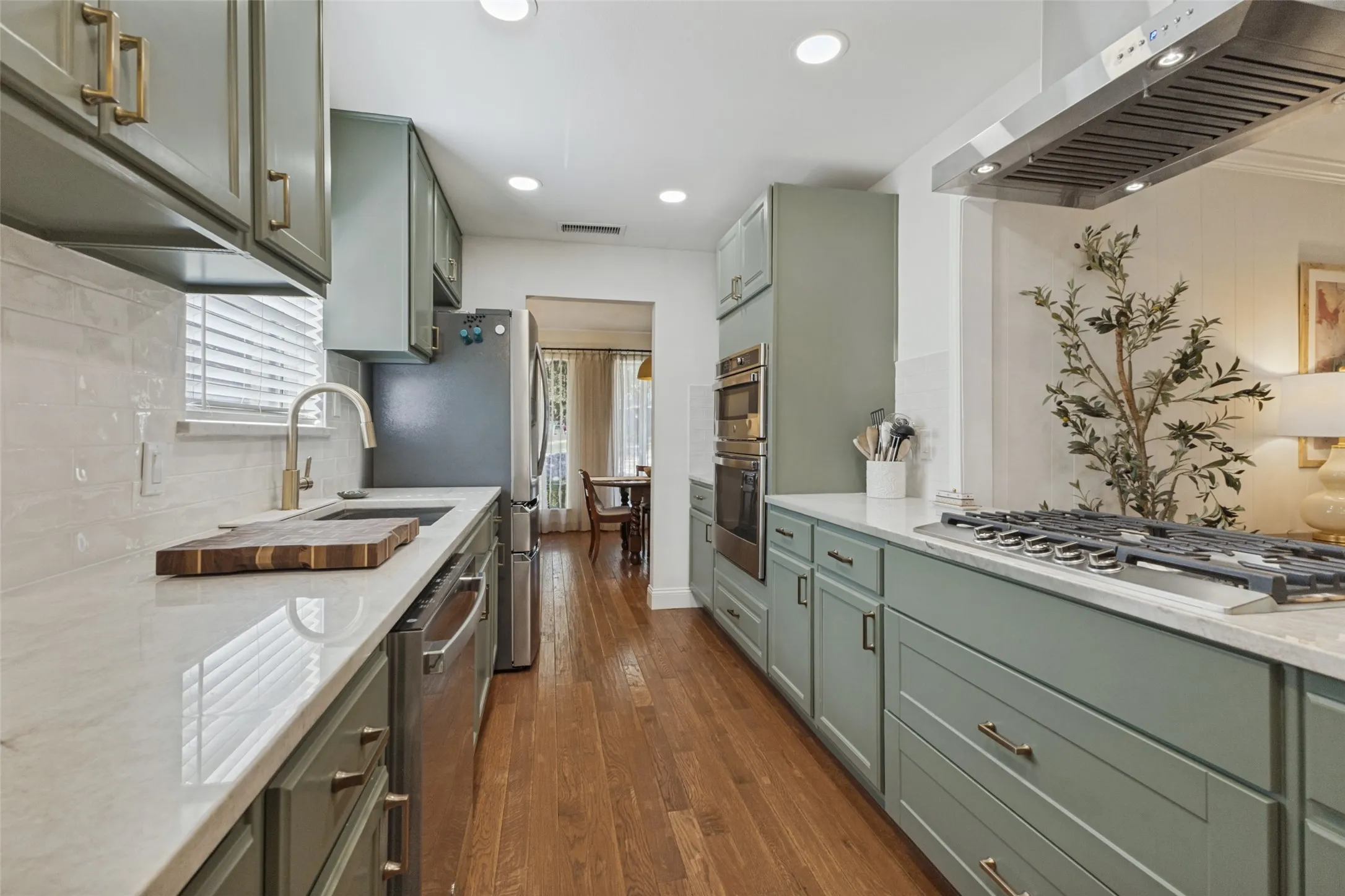 Galley kitchen featuring light green cabinetry, integrated stainless steel appliances, a gas range with a stainless steel vent hood, and wood-finish flooring