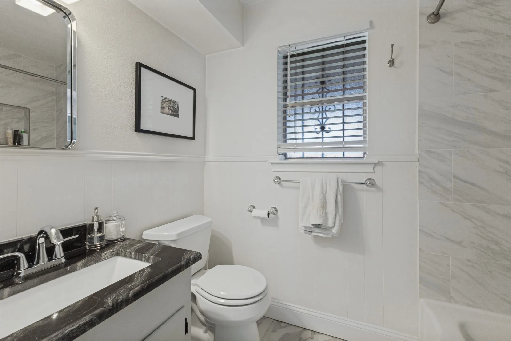 Vanity with dark countertop and undermount sink, wainscoting, and tiled shower surround