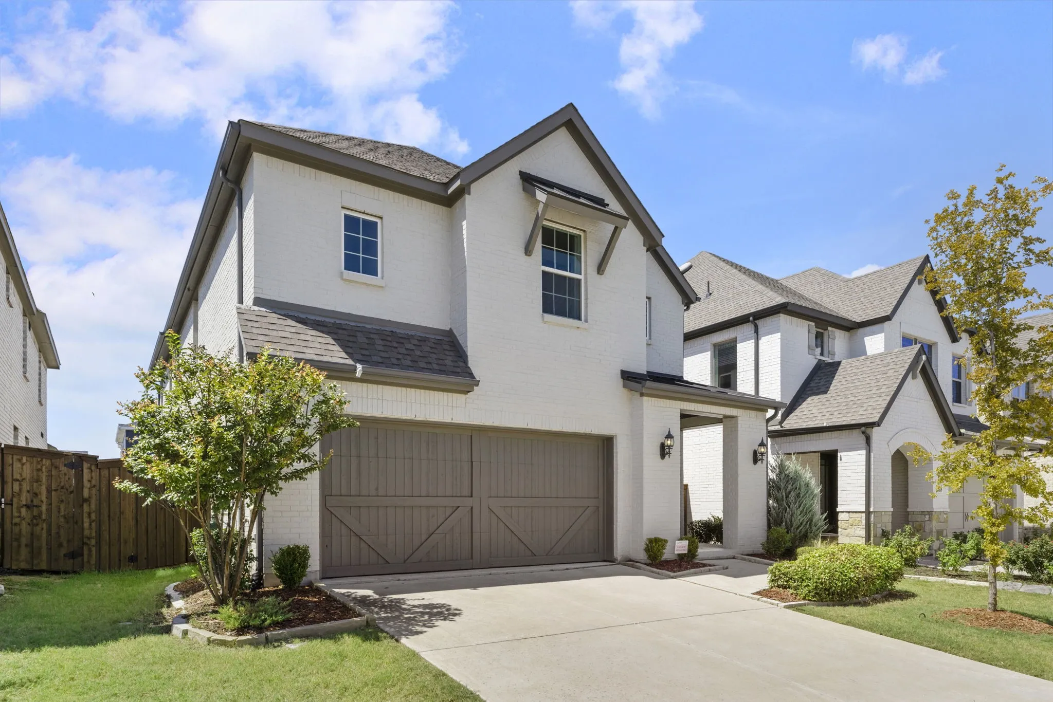 White brick exterior with dark trim accents, concrete driveway, two-car garage with wood-finish doors, and mature landscaping