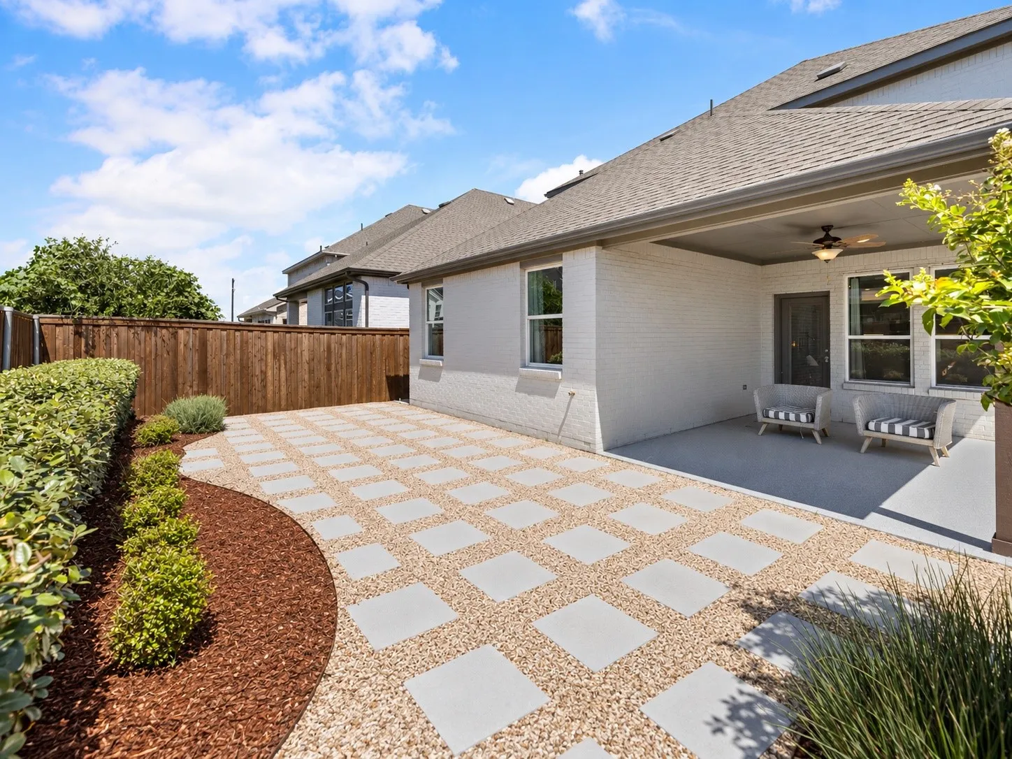 Expansive outdoor patio featuring paver and gravel hardscaping, a covered porch with a ceiling fan, a private wood fence, and landscaped borders with mature greenery