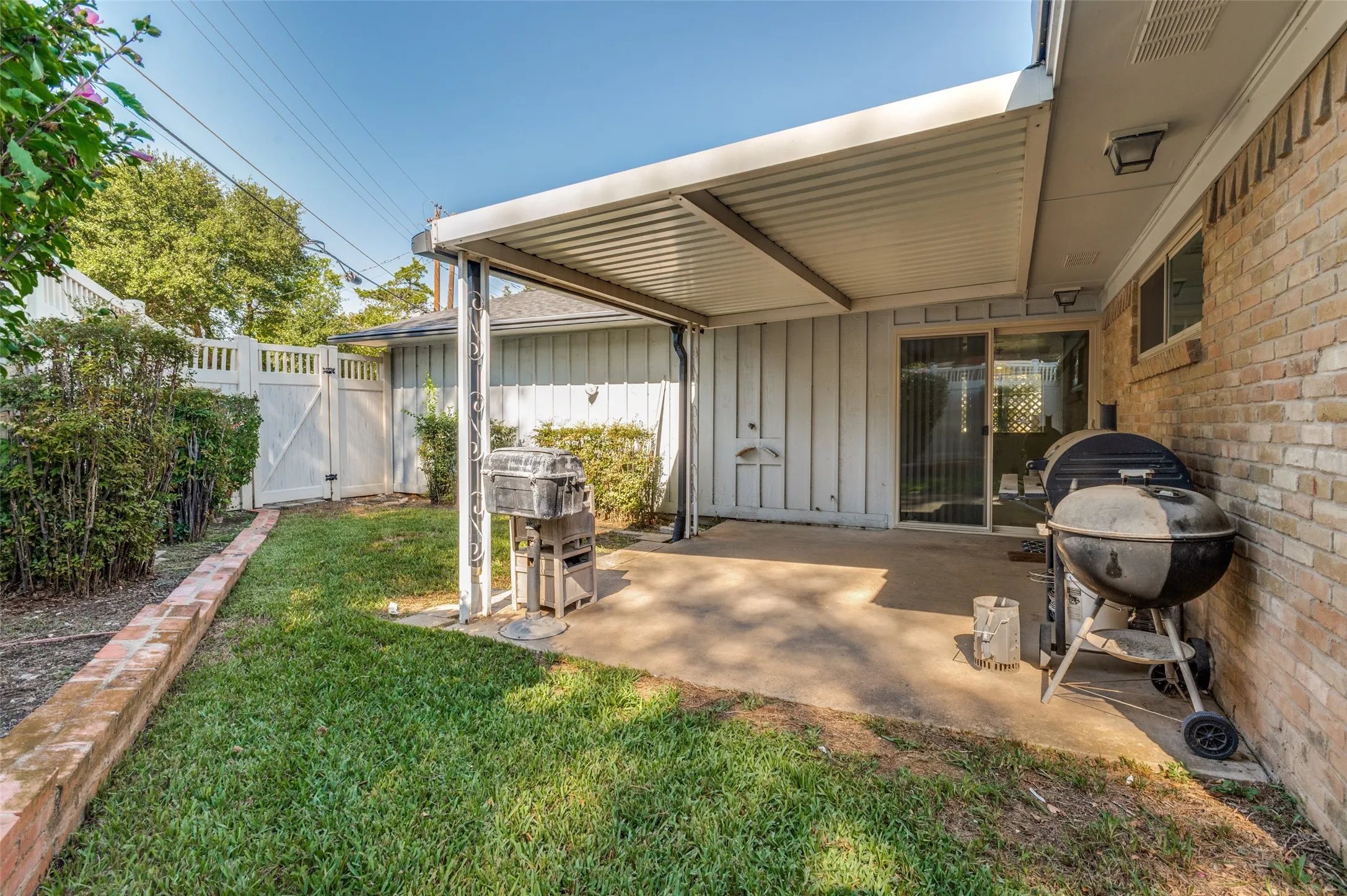 Expansive concrete patio with an attached covered awning, brick exterior, sliding glass door, and a white privacy fence with gate
