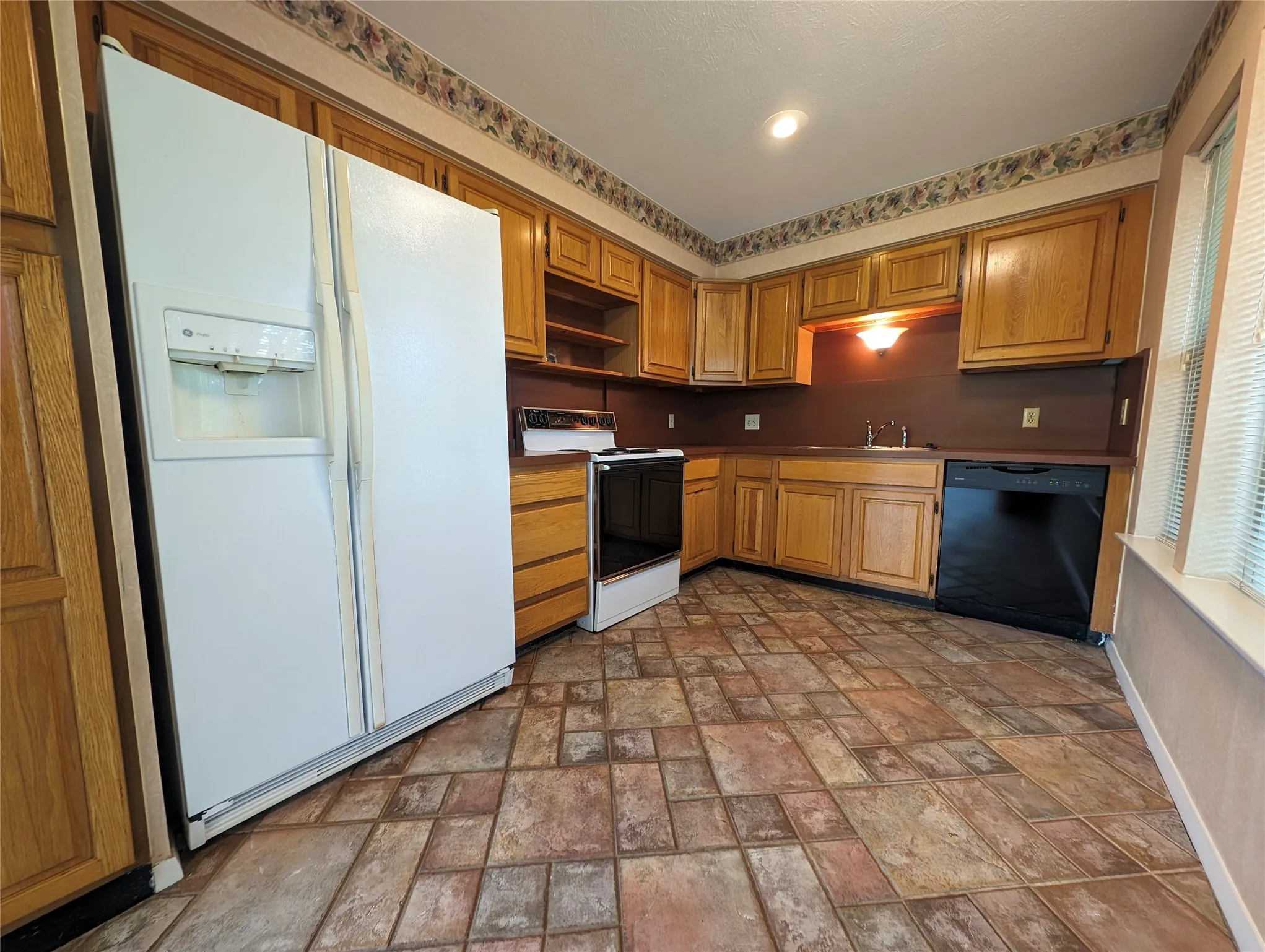 Kitchen featuring wood-finish cabinetry, built-in black dishwasher, white side-by-side refrigerator, under-cabinet lighting, and patterned tile flooring