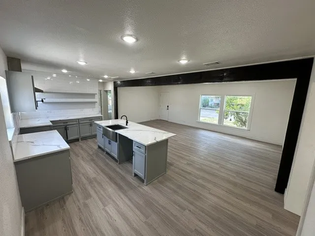 Wood-finish flooring throughout, recessed lighting, kitchen island with an integrated farmhouse sink and matte black faucet, light grey cabinetry, and white countertops with subtle veining