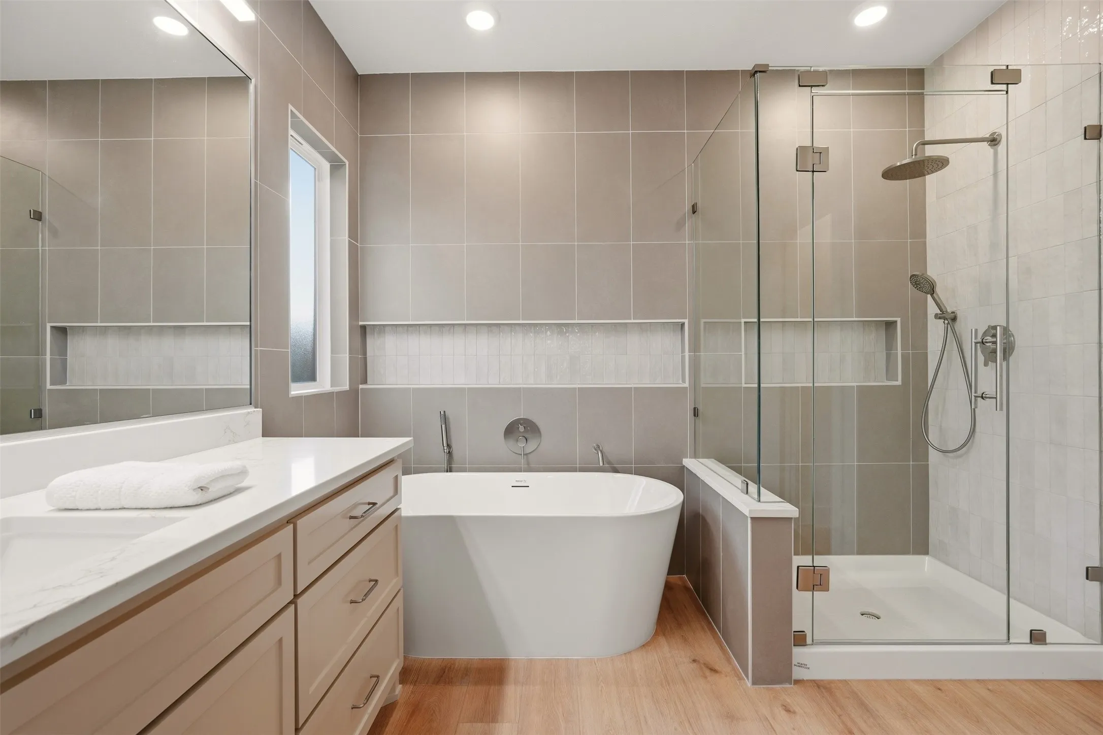 Walk-in shower with clear glass enclosure and rainfall showerhead, freestanding soaking tub, wood-finish flooring, vanity with light-colored countertop and drawers, and recessed wall niches