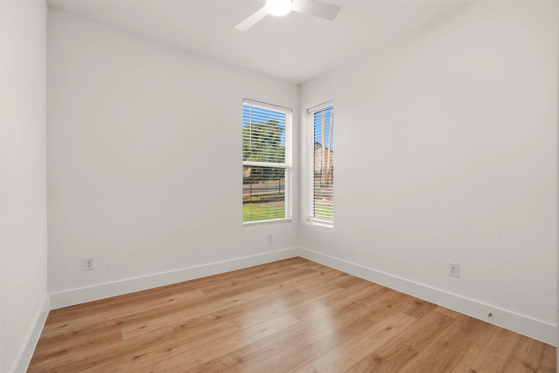 Wood-finish flooring, white walls, multiple windows with blinds, and an overhead ceiling fan