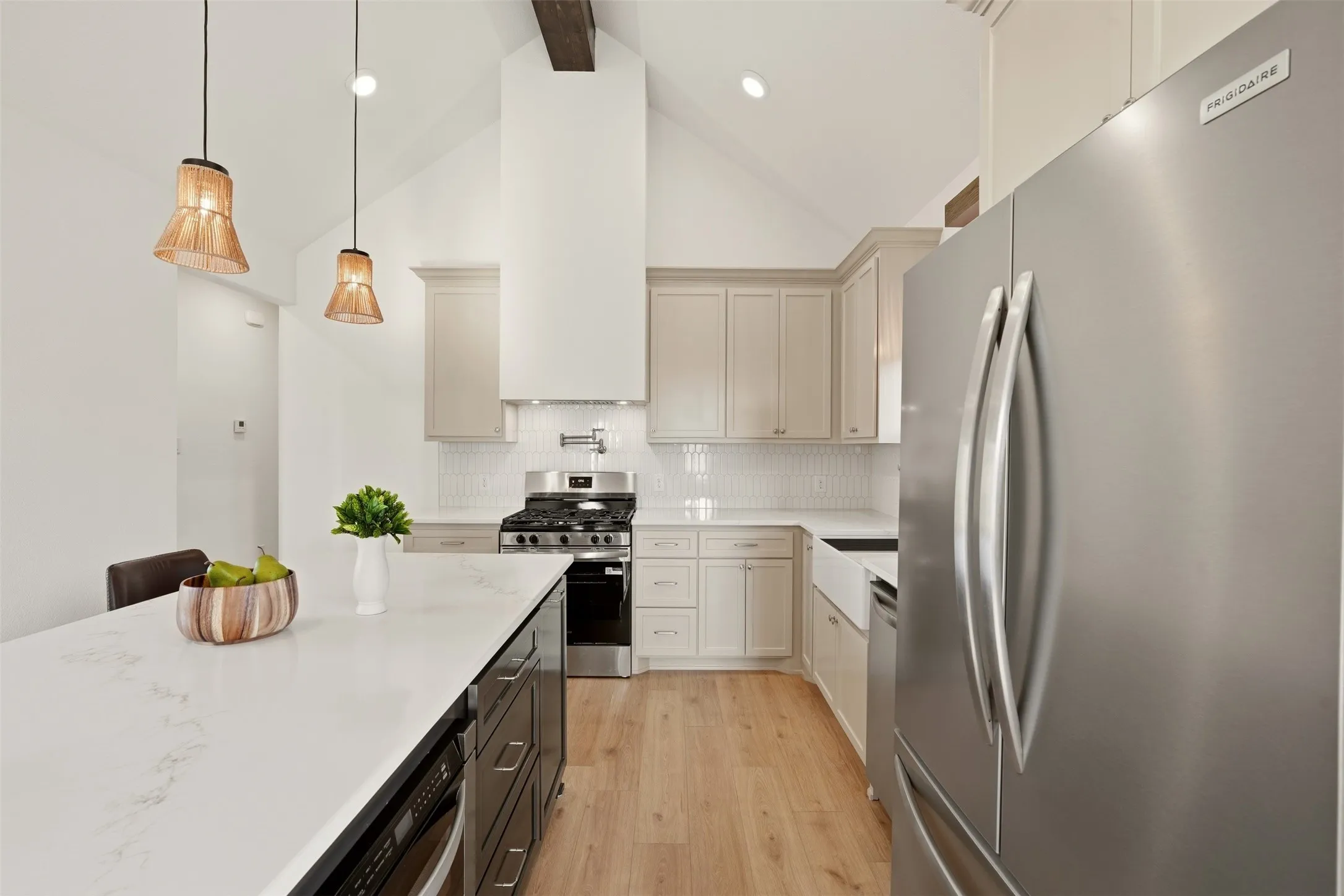 Vaulted ceiling with exposed beam, stainless steel appliances, white subway tile backsplash, farmhouse-style sink, and light wood-finish flooring