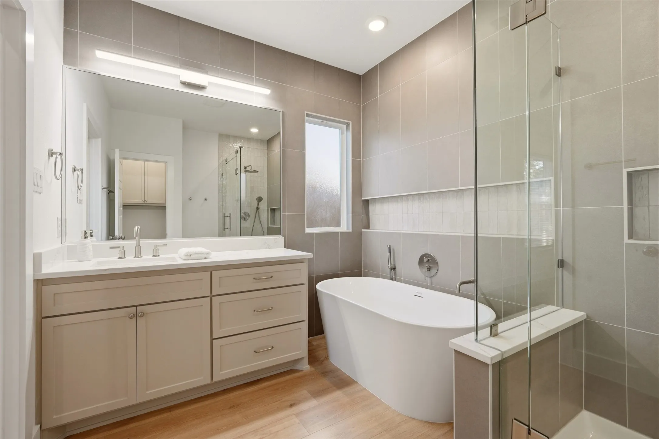 Freestanding soaking tub, frameless glass shower enclosure with built-in bench, light wood-finish flooring, vanity with light-colored countertop, and recessed lighting