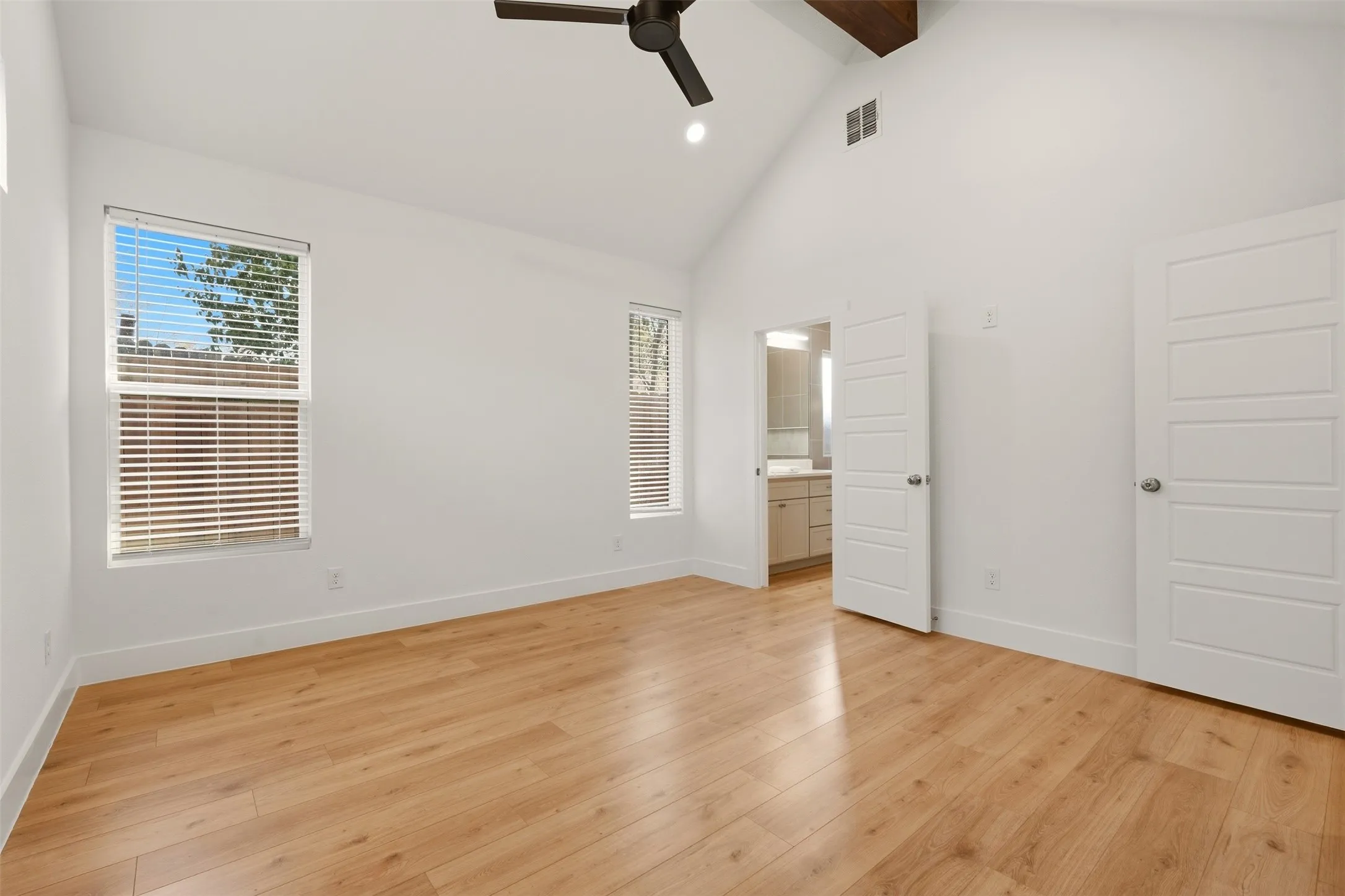Vaulted ceiling with exposed wood beam, wood-finish flooring, two windows with blinds, ceiling fan, and white panel doors