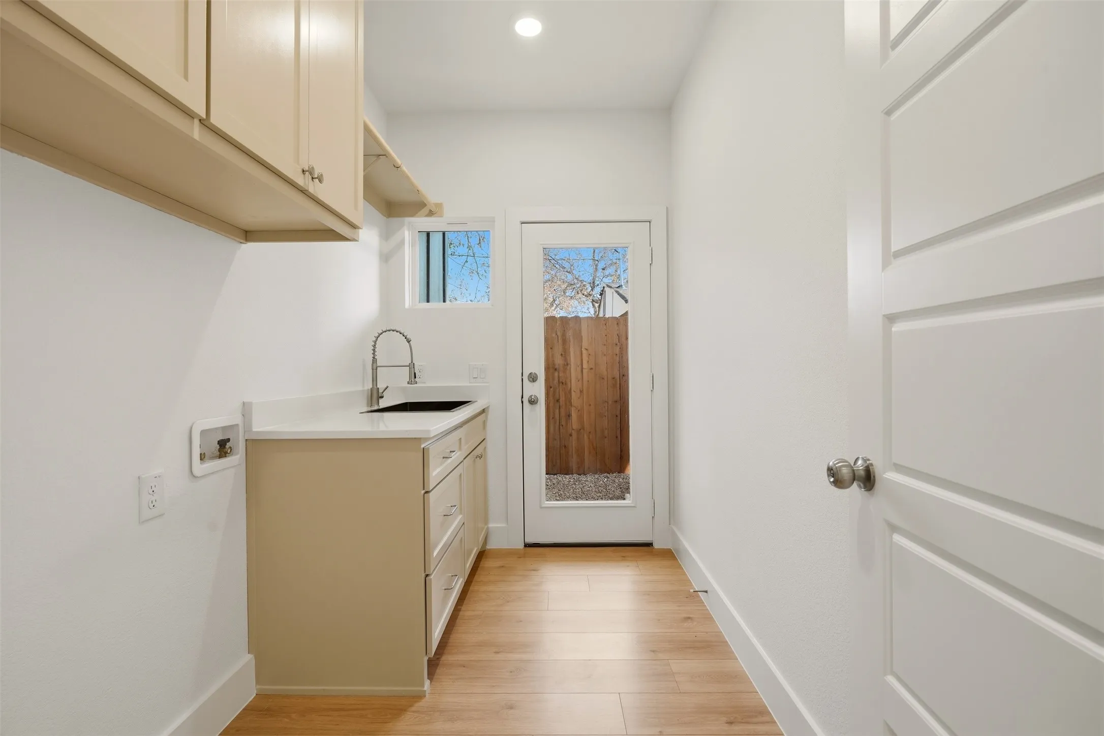 Laundry area featuring wood-finish flooring, light-toned cabinetry, a built-in sink with a gooseneck faucet, a light-toned countertop, and a glass-paneled exterior door