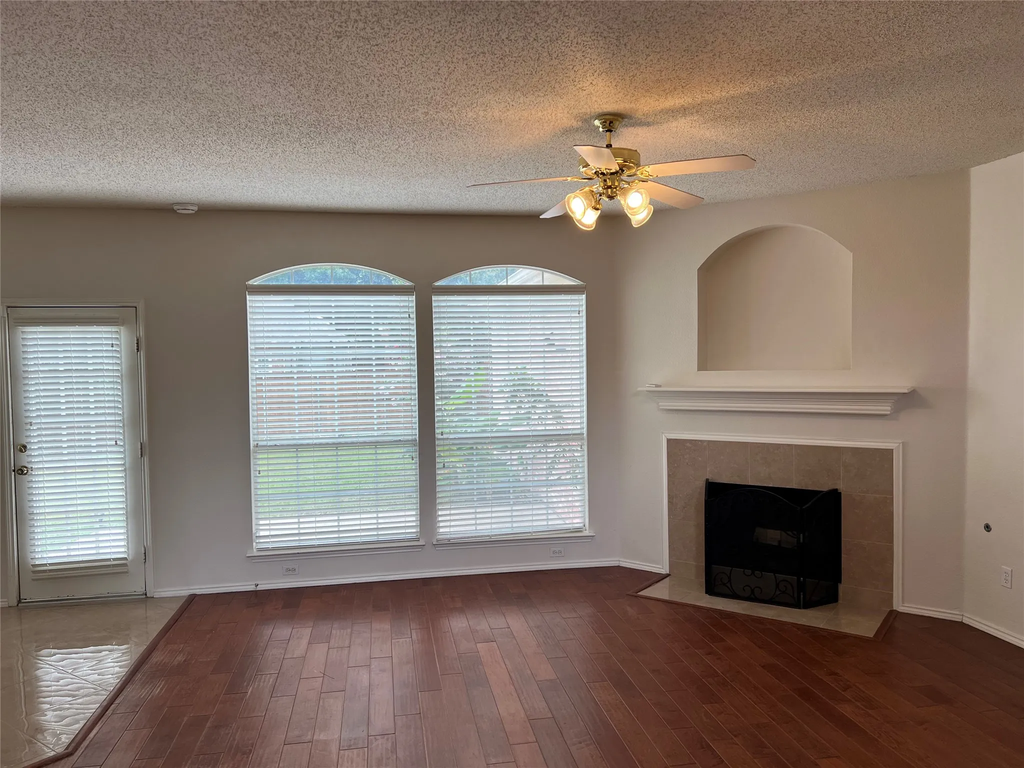 Wood-finish flooring, tiled fireplace with mantel, arched wall niche, multiple windows, and a ceiling fan with integrated lighting