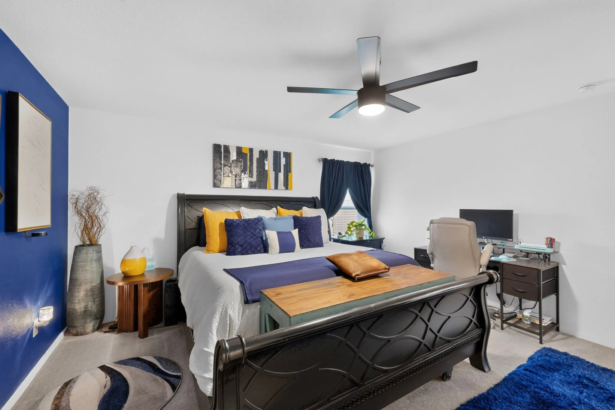 Primary Bedroom Suite featuring a black metal platform bed frame, crisp white and navy bedding accents with gold throw pillows, ceiling-mounted fan with integrated lighting, dark wood nightstands, and soft area rug