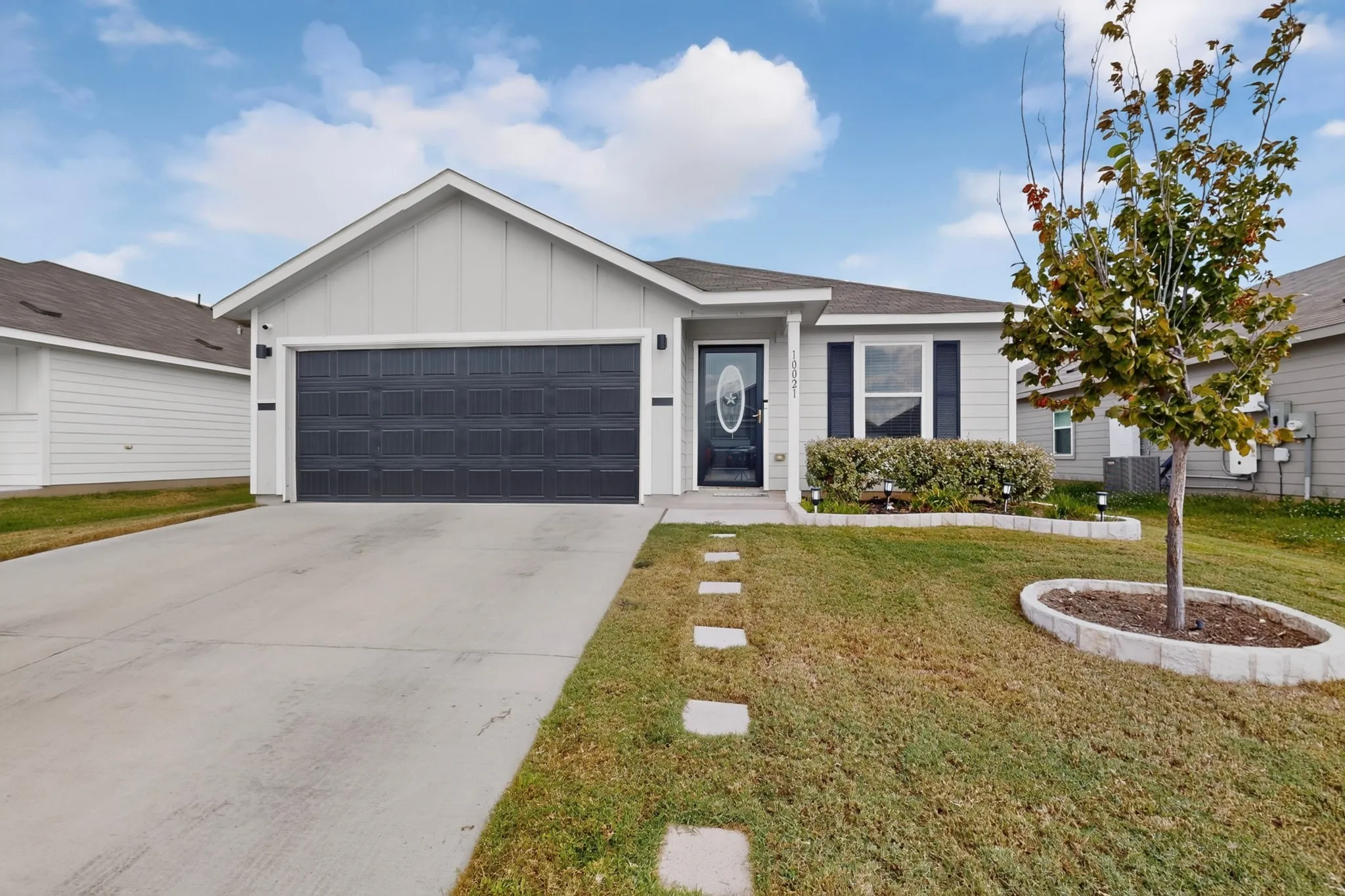 Light-colored siding, dark garage door, concrete driveway, stone paver pathway, and established front landscaping