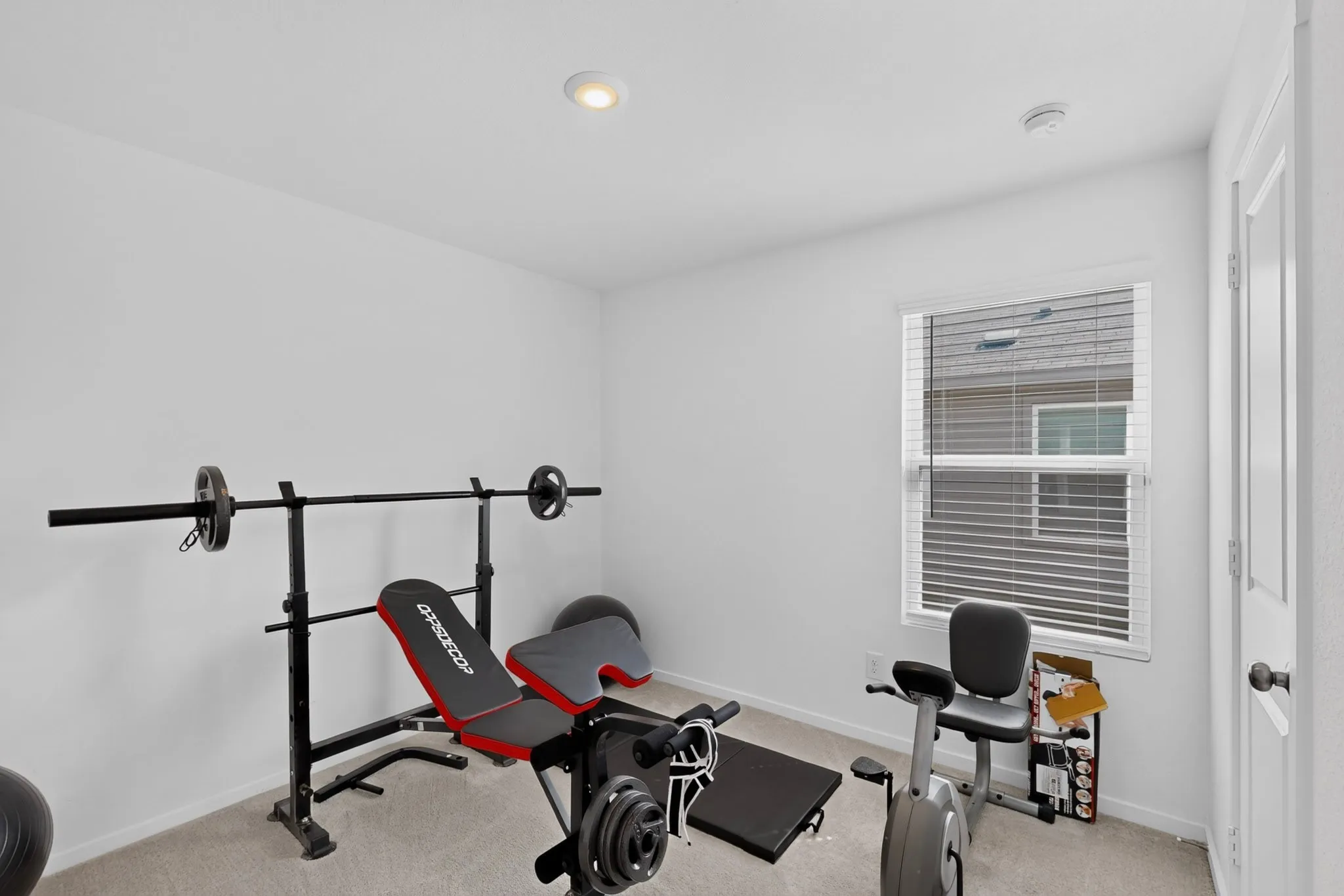 Recessed lighting illuminates a room with carpet flooring, a single window with blinds, and a white paneled door