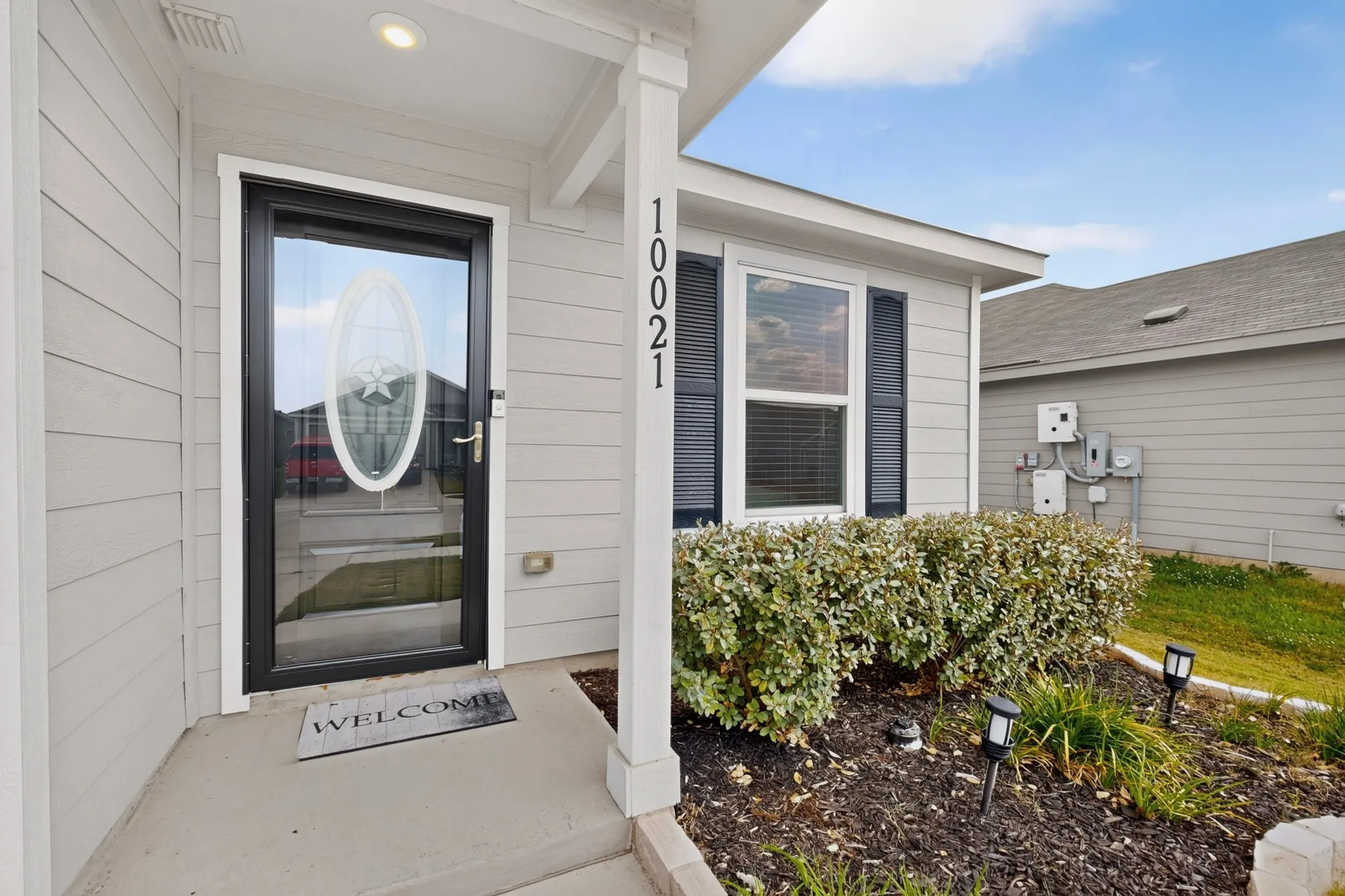Welcoming entry featuring a black-framed glass storm door with oval window insert, white horizontal siding, recessed ceiling lighting, and a manicured landscape bed with established shrubs and fresh mulch
