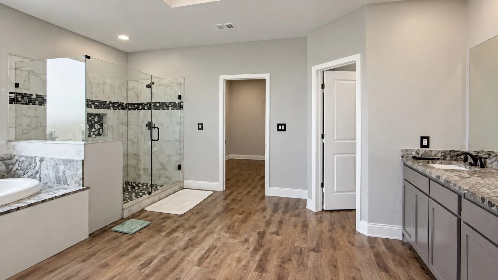 Spacious bathroom featuring wood-finish flooring, a frameless glass shower enclosure, double vanity with stone countertops, and recessed lighting