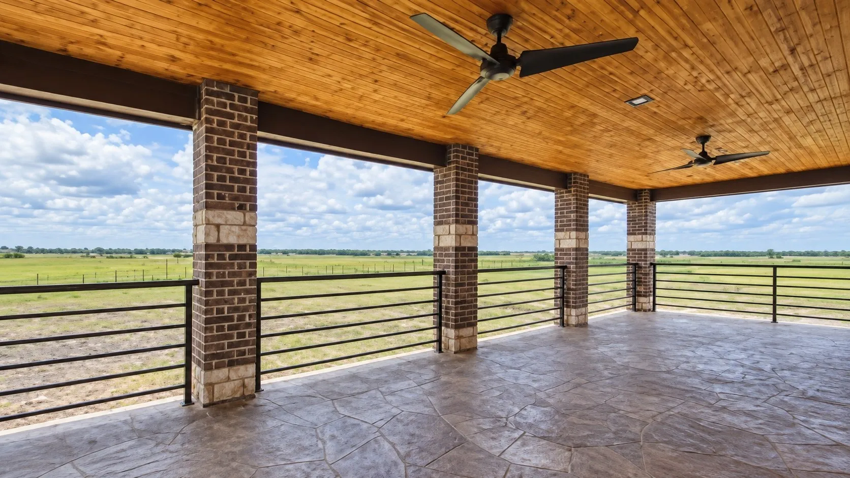 Expansive covered patio featuring a wood-finish ceiling, dual ceiling fans, brick and stone columns, black metal railings, and a patterned concrete floor