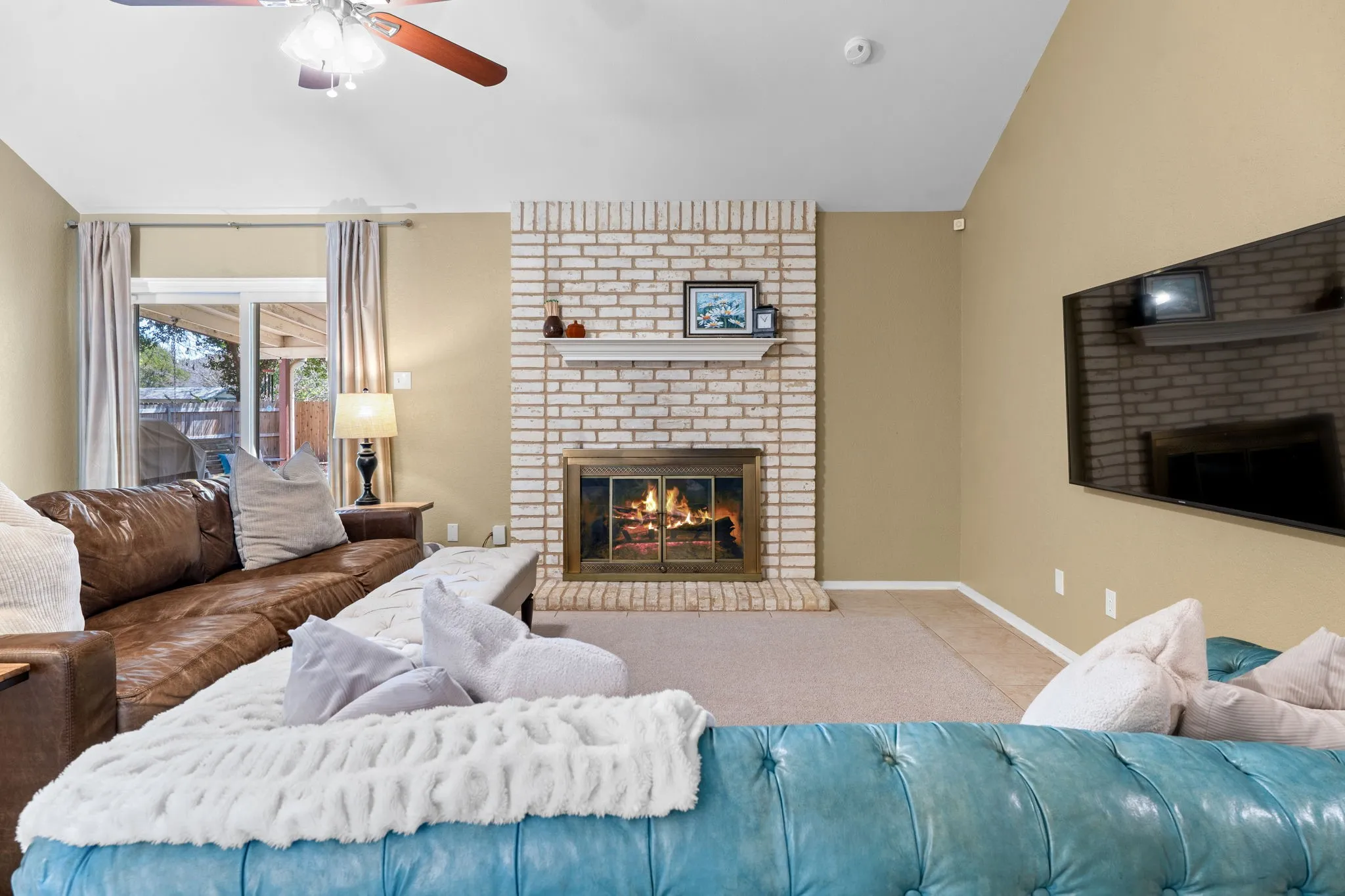 Vaulted ceiling, ceiling fan with integrated lighting, brick fireplace with mantel, glass-paneled fireplace insert, and sliding glass door