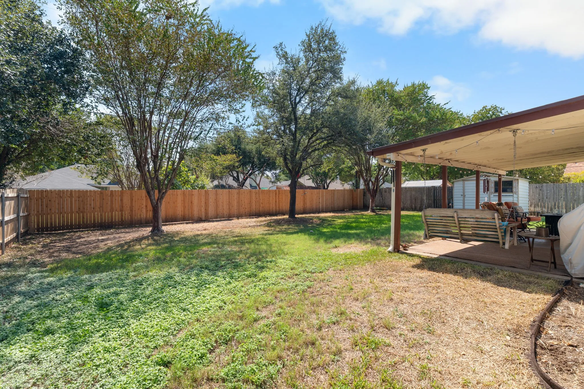 Expansive backyard oasis featuring mature shade trees, lush lawn, and a covered patio structure with wood-beam construction