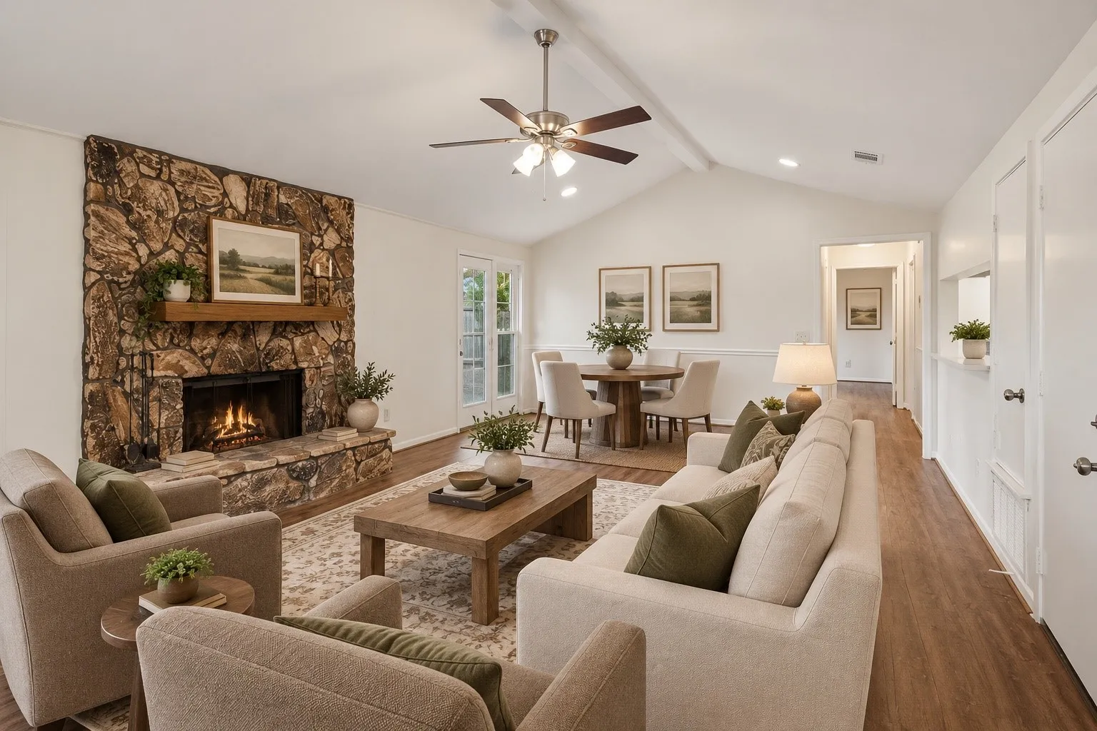 Spacious open-concept living area featuring a wood-burning fireplace with stone surround, hardwood flooring throughout, and vaulted ceilings with ceiling fan