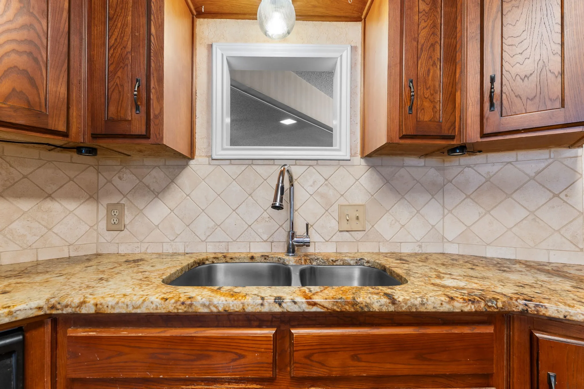 Granite countertops, stainless steel double basin sink, chrome gooseneck faucet, diamond-patterned tile backsplash, and wood cabinetry