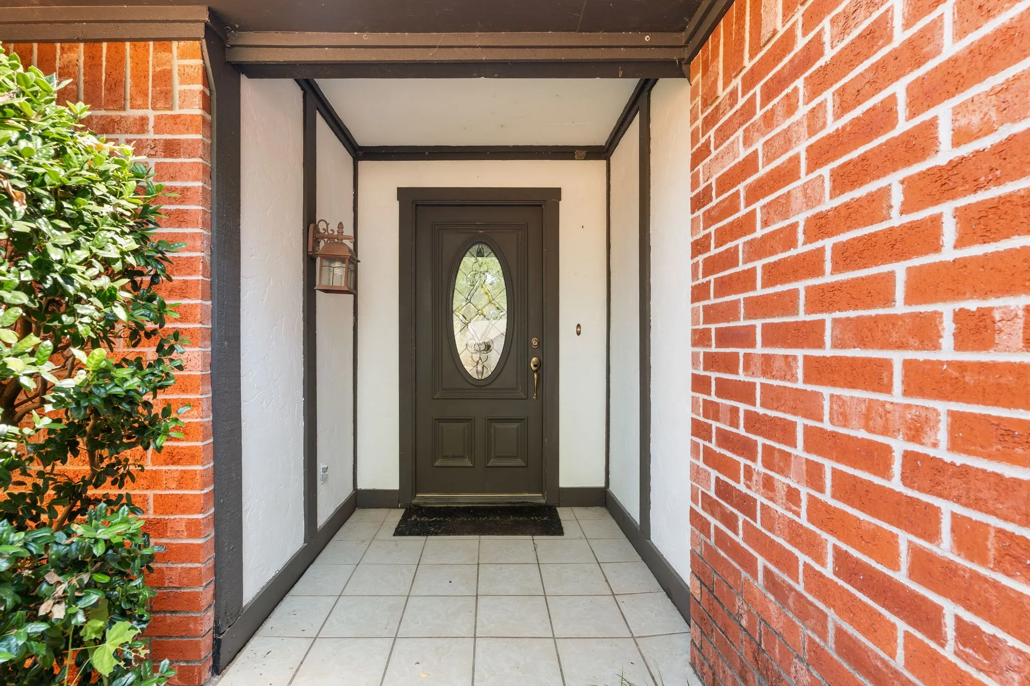 Brick exterior walls with white mortar, recessed entry, dark wood-finish entry door with oval glass insert, exterior wall light fixture, and light-colored tile flooring