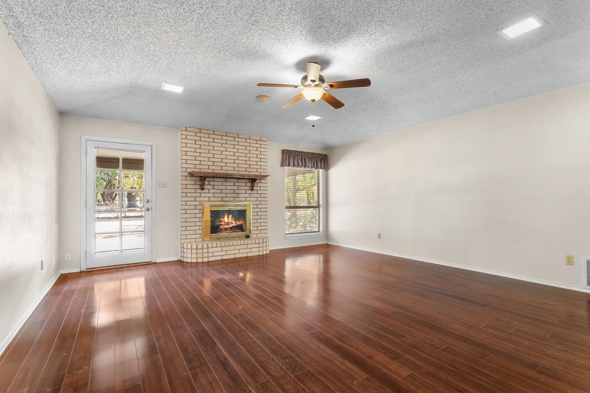 Wood-finish flooring, brick fireplace with a wood mantle, ceiling fan, and a glass-paneled door