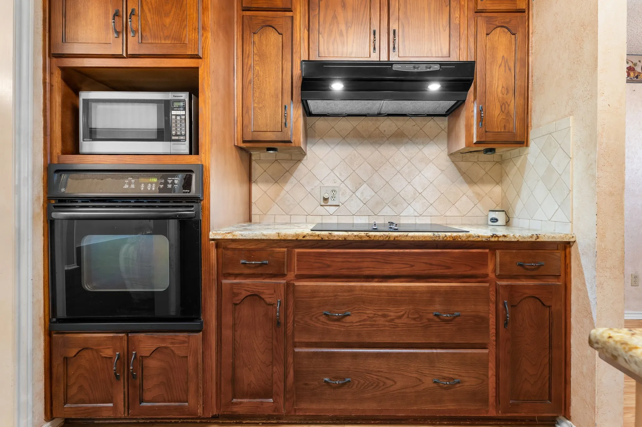 Rich wood cabinetry with dark bronze hardware, built-in oven, built-in microwave, integrated cooktop, and speckled stone countertops