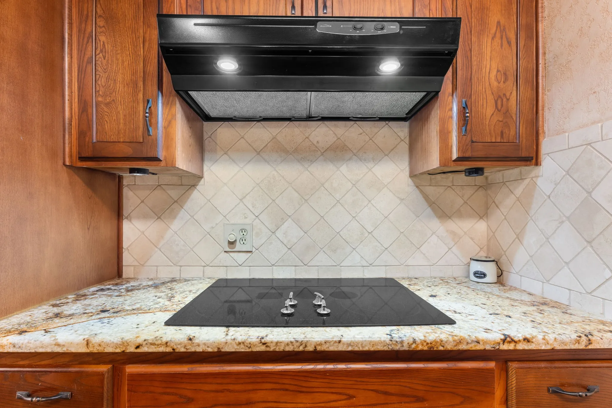 Built-in cooktop with a range hood, natural wood cabinetry, light-toned stone countertops, and a diagonal tile backsplash