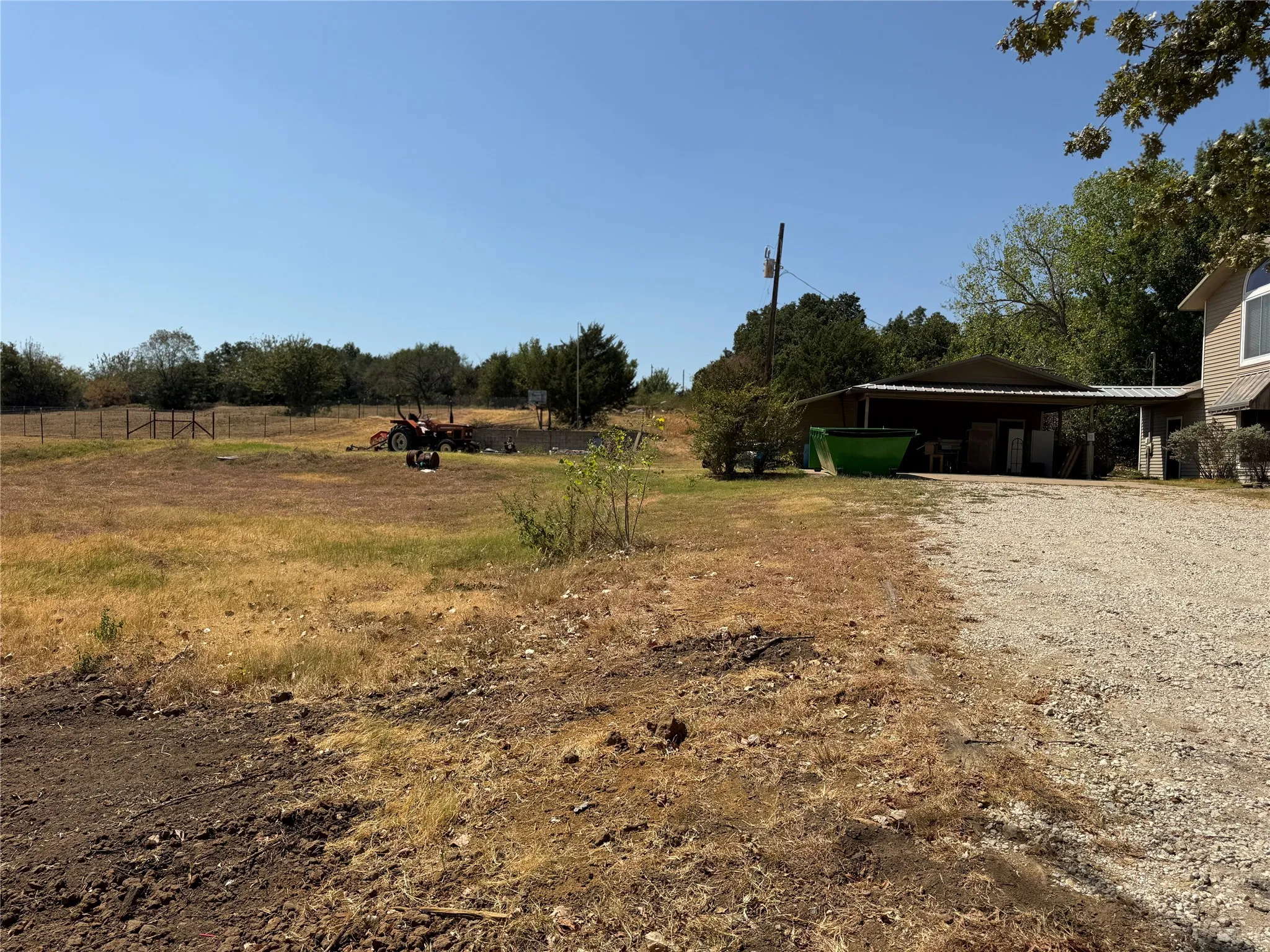 Gravel driveway with a metal carport structure, mature trees, and an expansive open field