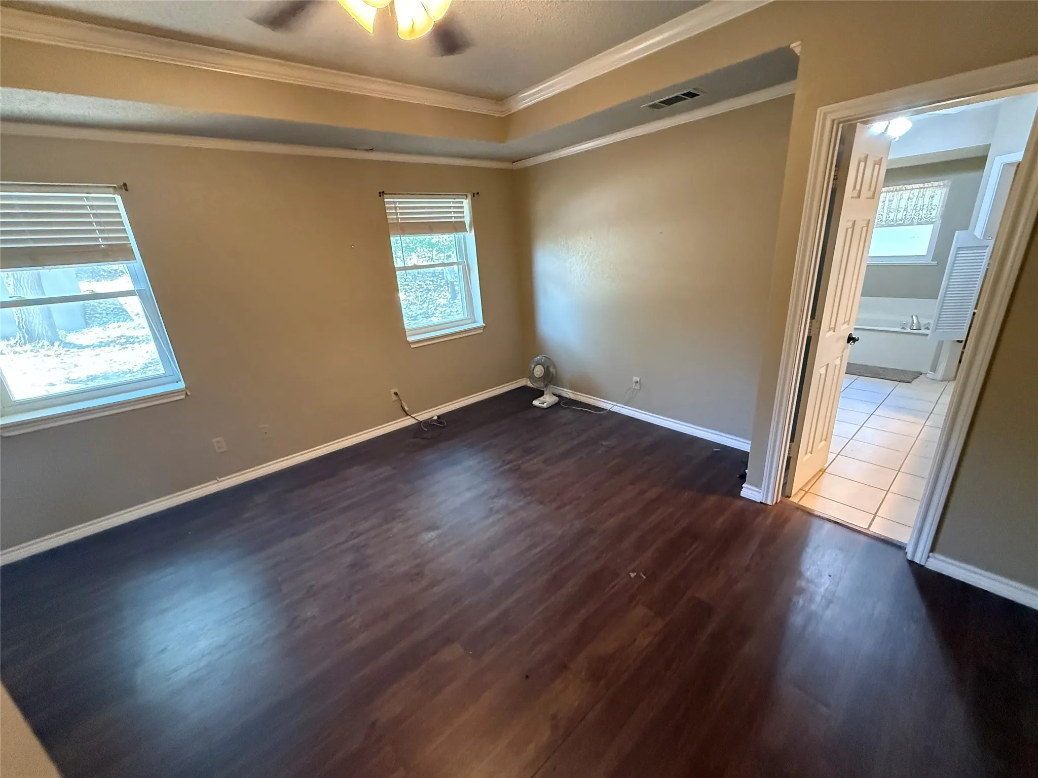 Tray ceiling with crown molding, wood-finish flooring, two windows with blinds, ceiling fan with integrated lighting, and an open doorway