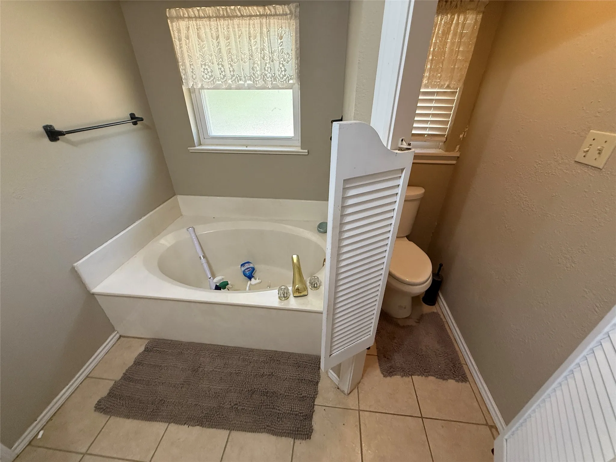 Built-in corner bathtub, tiled flooring, louvered privacy partition, wall-mounted towel bar, and a frosted window