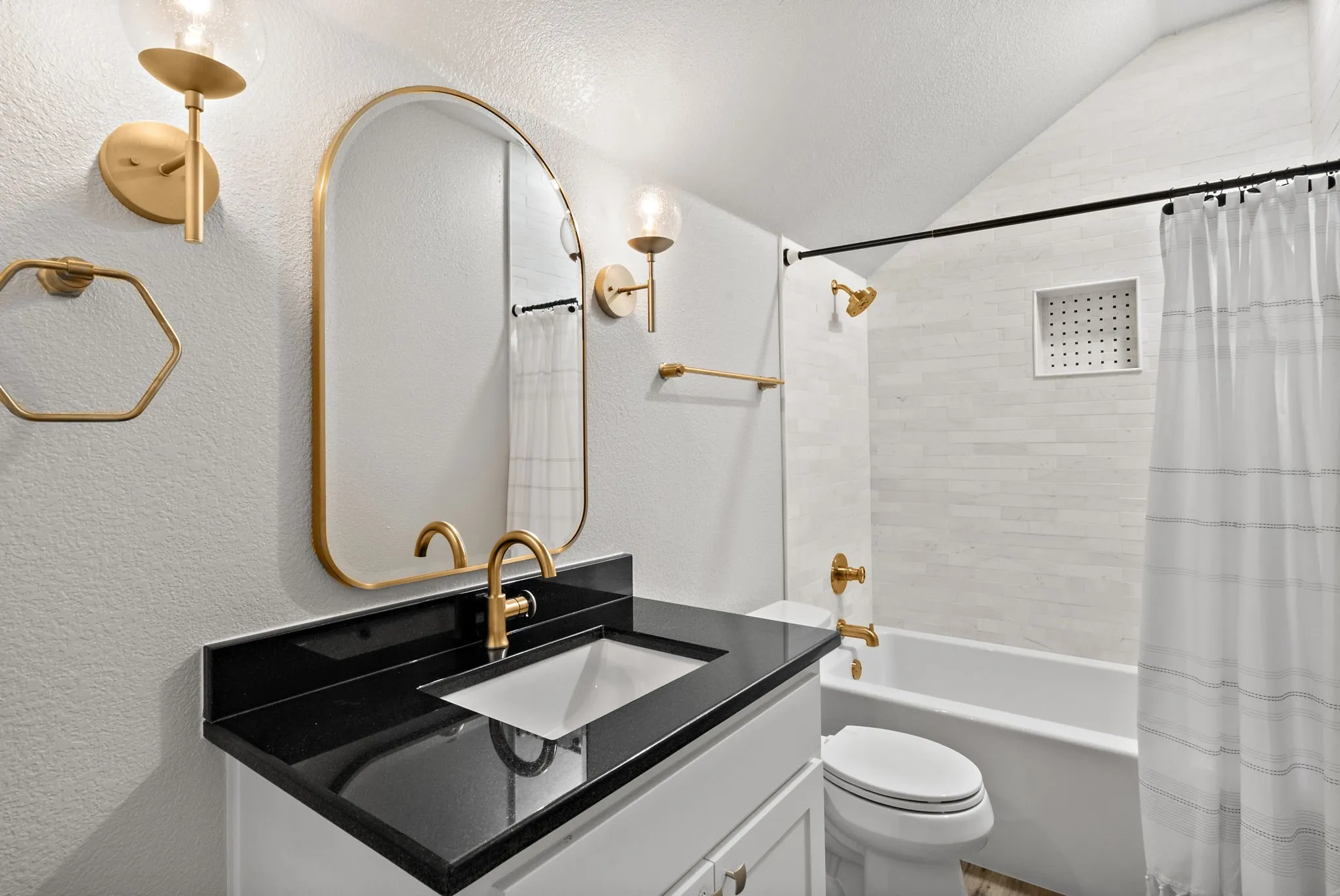 Vanity with a black countertop and integrated sink, gold-finish fixtures, white subway tile tub surround with recessed niche, and a white bathtub