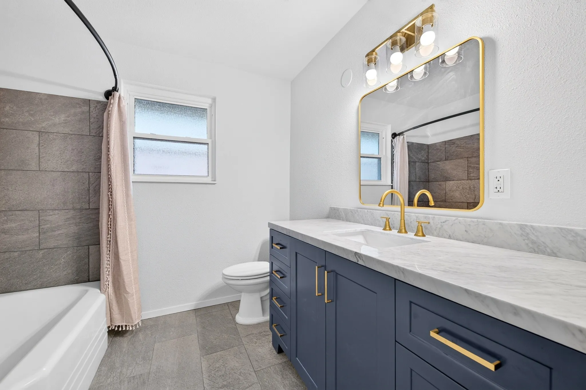 Bathroom featuring a white bathtub, gray tiled shower surround, dual-sink vanity with blue cabinetry, gold-finish hardware, and a framed mirror with integrated lighting