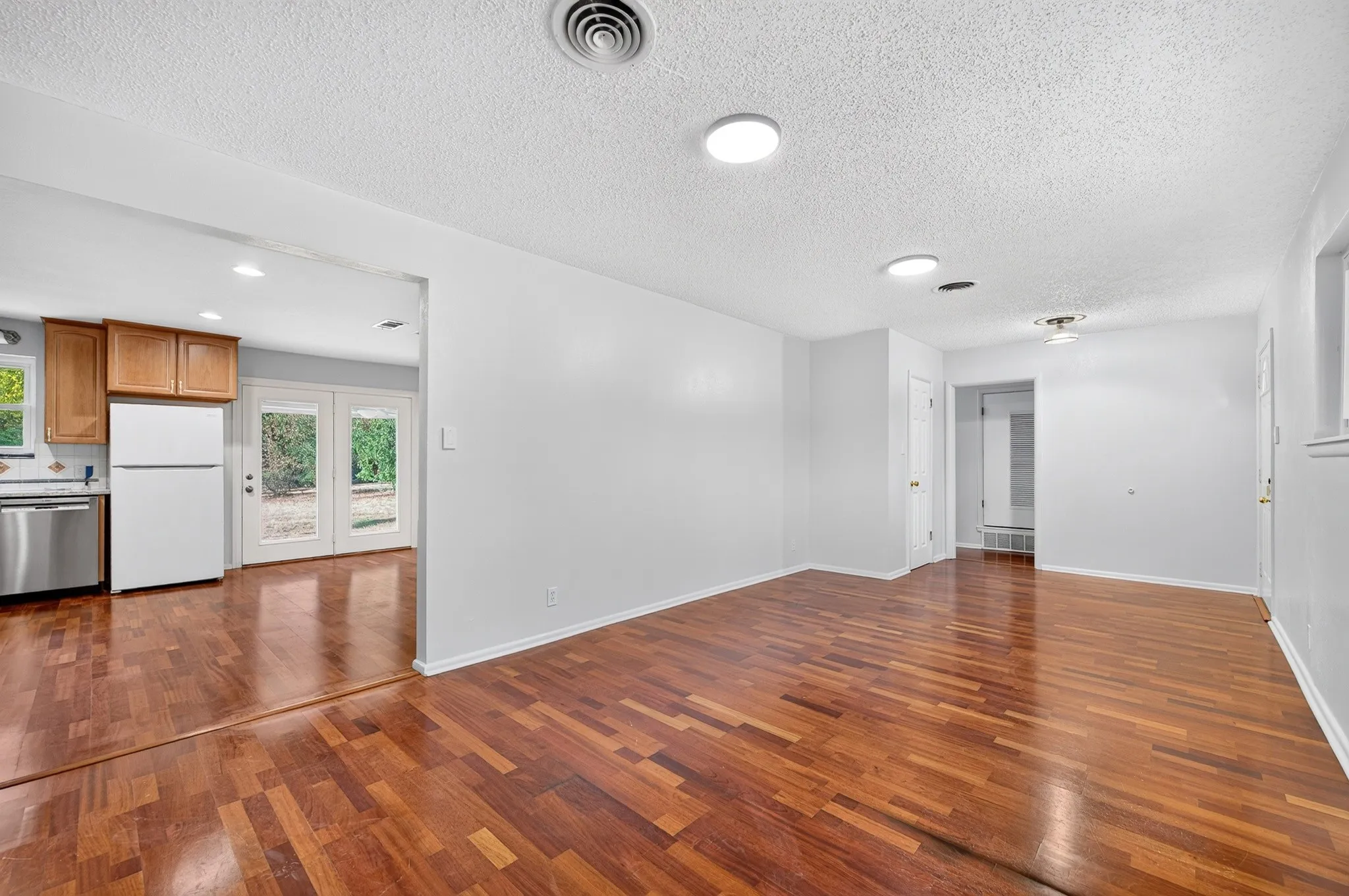 Spacious open-concept living area showcasing rich wood-finish flooring throughout, abundant natural light from multiple windows, recessed ceiling lighting, and seamless access to the kitchen featuring stainless steel appliances and white cabinetry