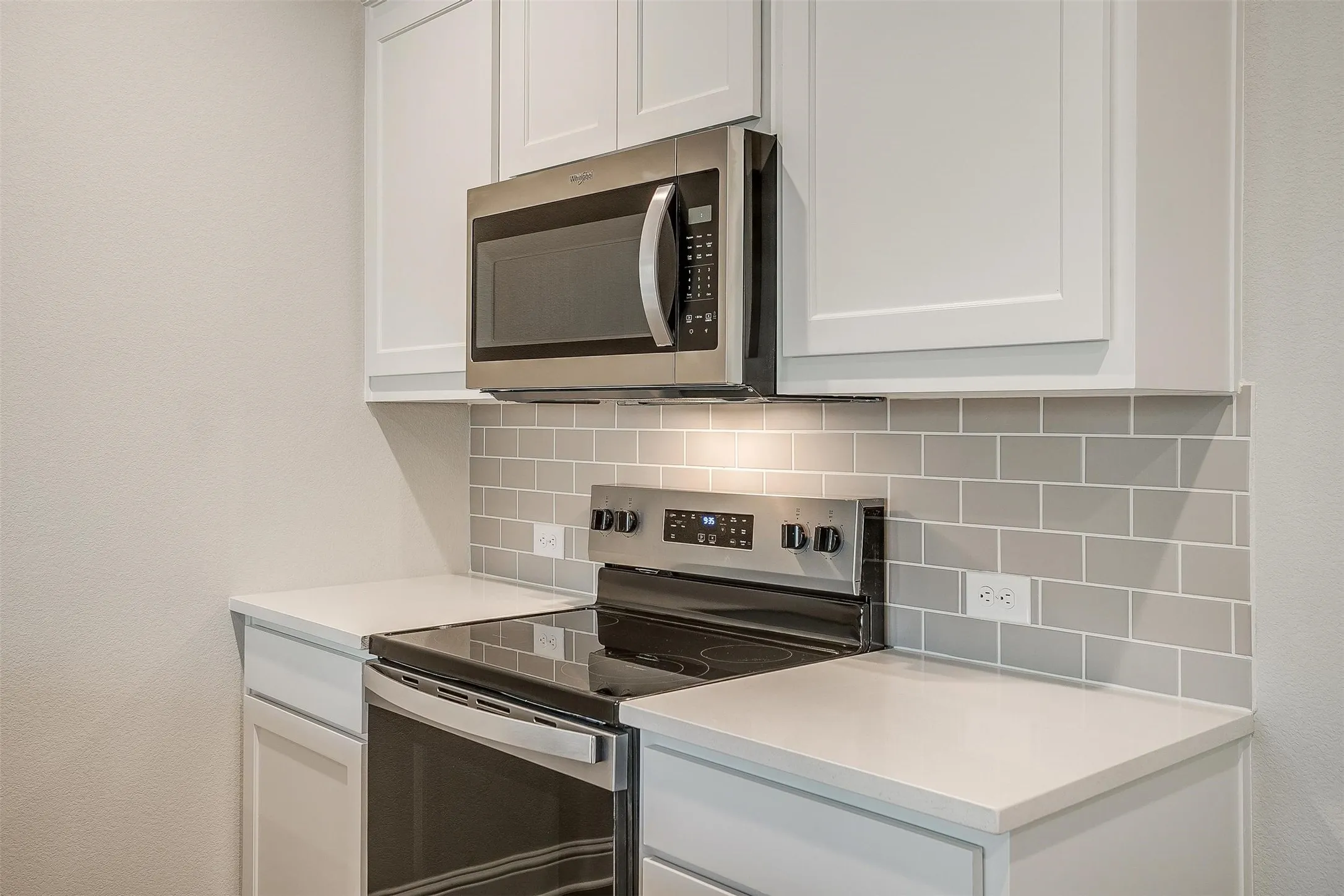 Stainless steel appliances with an electric range, built-in microwave, light gray subway tile backsplash, white shaker-style cabinetry, and light-toned countertops