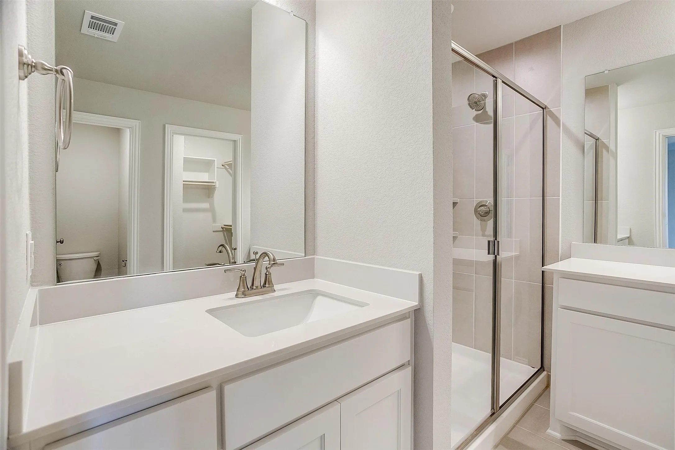 Built-in vanity with a single basin sink and brushed nickel faucet, large wall mirror, glass-enclosed shower with tile surround, and white cabinetry with solid countertops