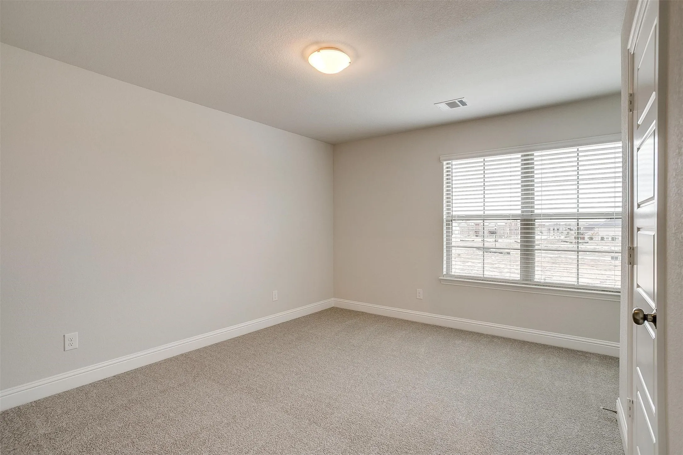 Neutral wall paint with textured finish, light-toned carpet, window with horizontal blinds, overhead flush mount lighting, and white trim